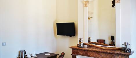 A hotel room with a wooden table, a chair, a television mounted on the wall, and a large mirror above a marble counter.