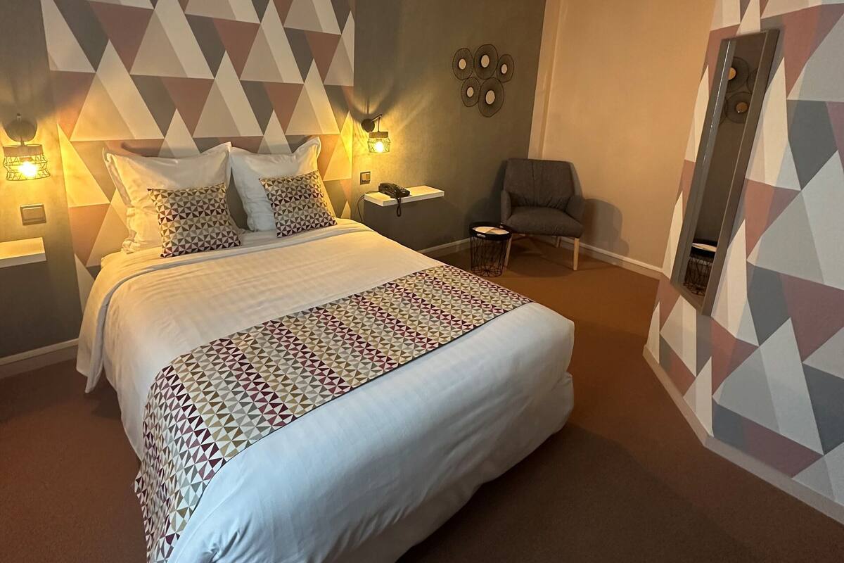 A hotel room with a large bed, a bedside table, a chair, and a wall with geometric patterns.