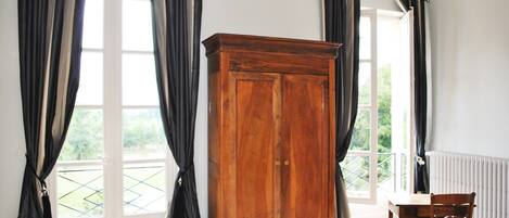 Una habitación con un armario de madera, una mesa de comedor con una silla y ventanas grandes con cortinas.