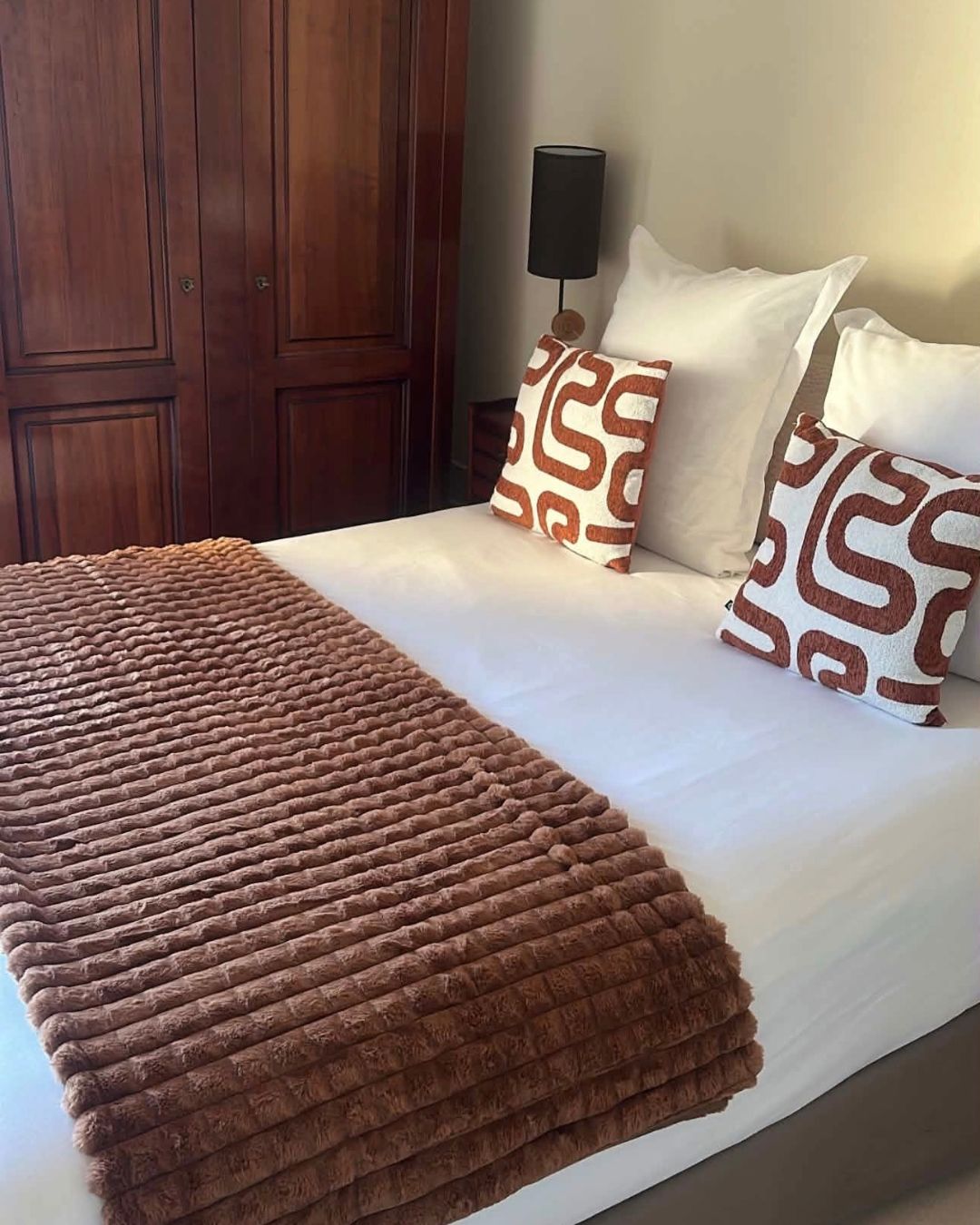 A bed with a brown blanket, two patterned pillows, and a bedside lamp.