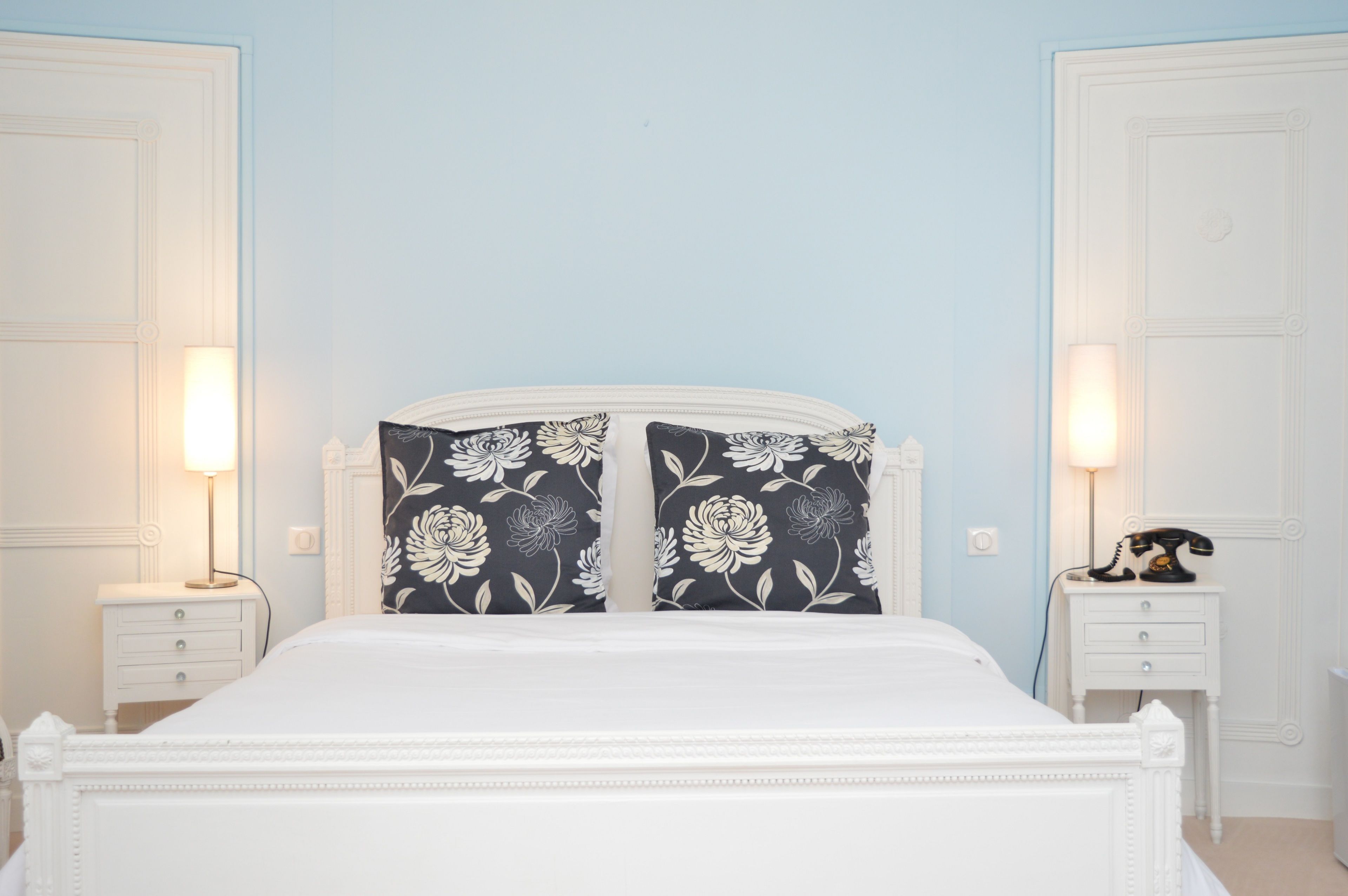 A bedroom with a white bed, floral-patterned pillows, two bedside tables with lamps, and a telephone on one of the tables.