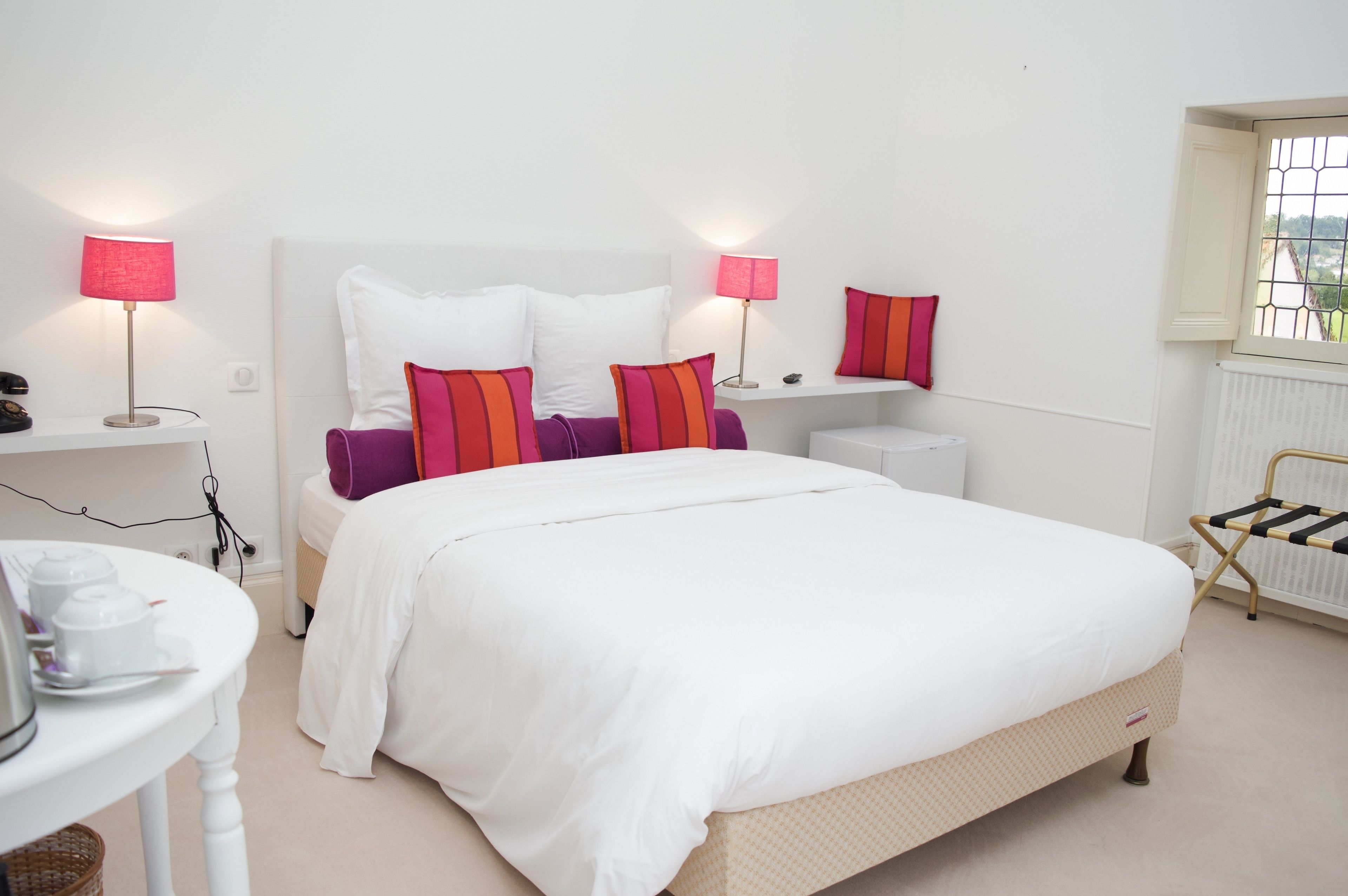 A neatly arranged bedroom with a large bed, bedside lamps, a small table with toiletries, and a window with a view.
