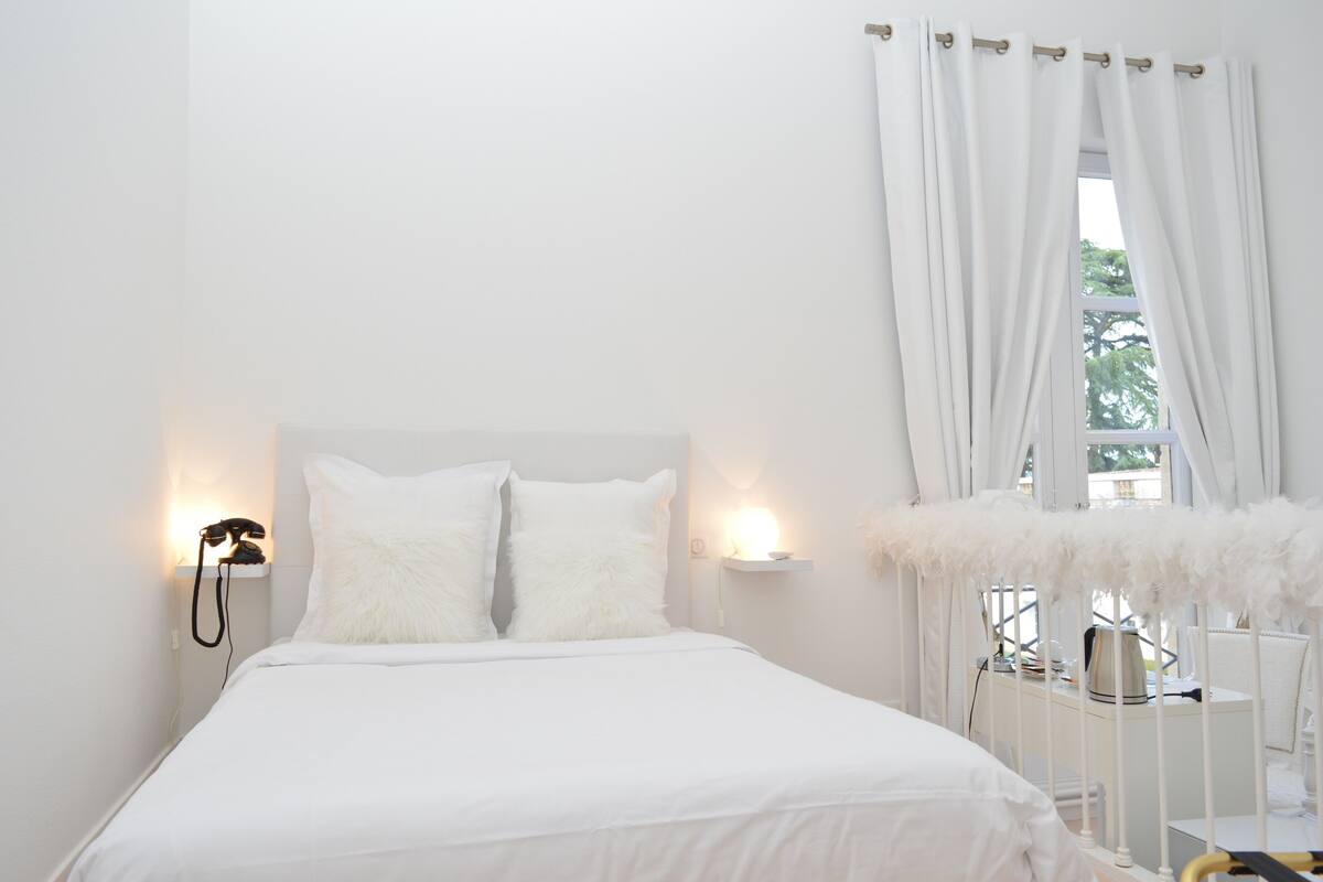 A white bedroom with a bed, two white pillows, a bedside lamp, a nightstand with a telephone, and a window with white curtains.