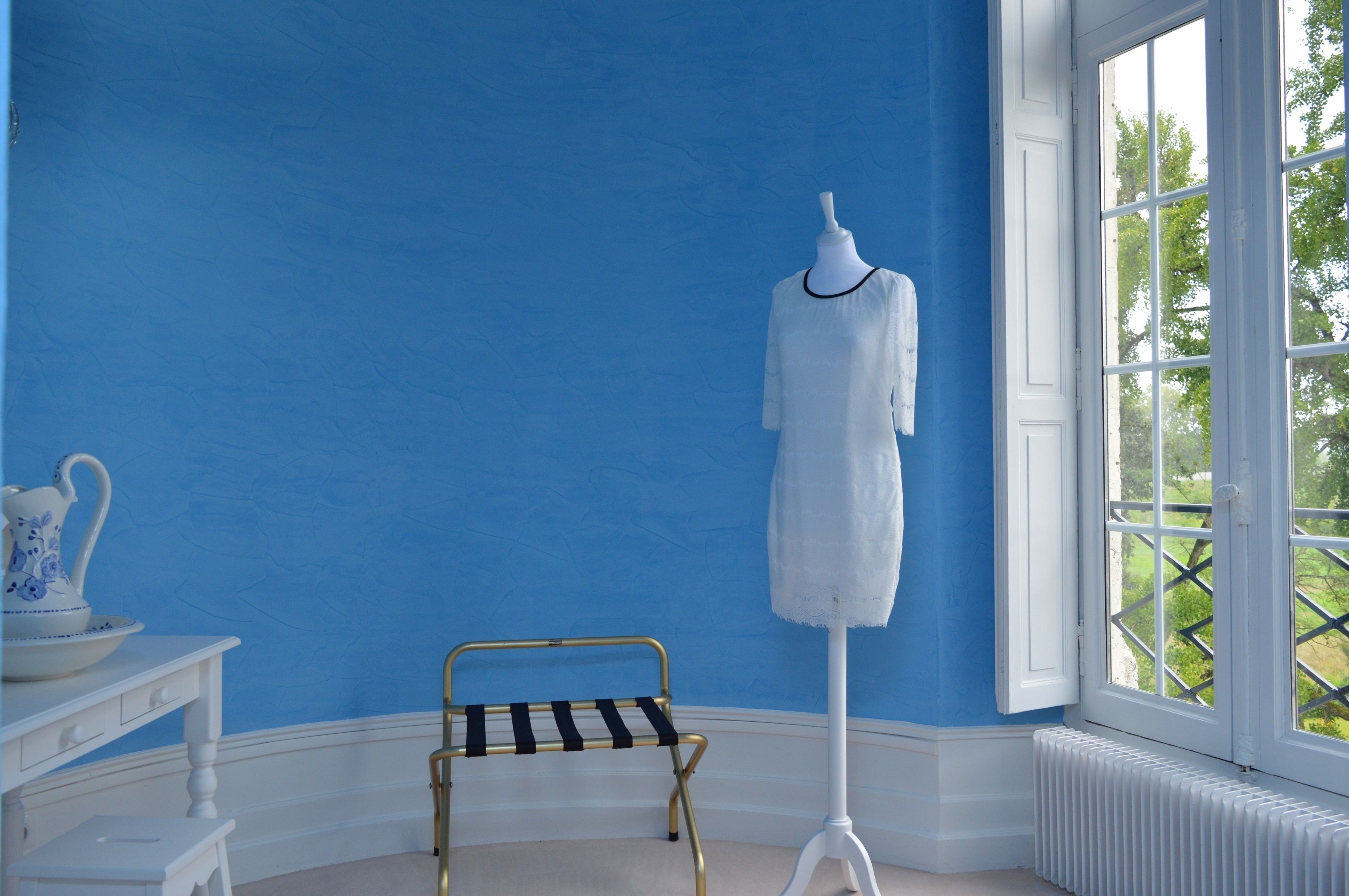 A room with a mannequin in a white dress, a small black and white striped chair, and a white radiator near a window with a view of greenery.