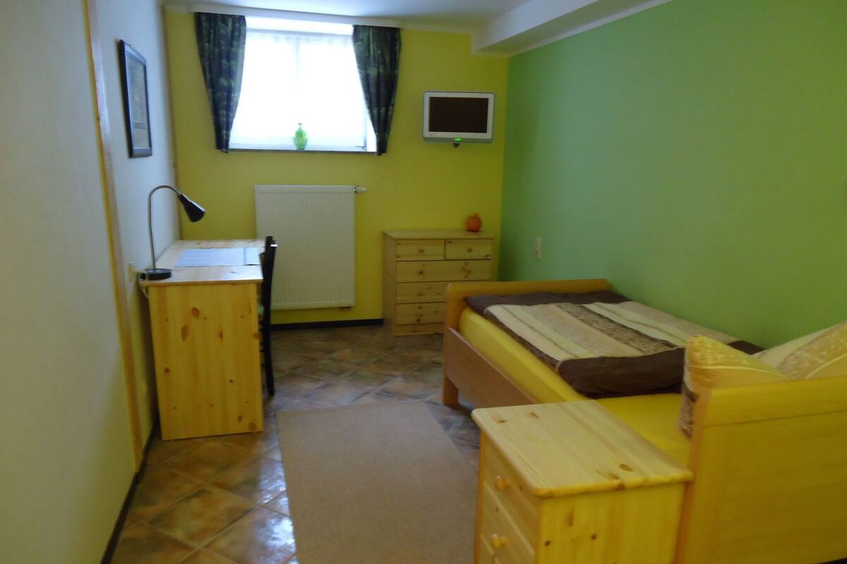 A small, brightly colored room with a bed, a desk, a dresser, and a window with curtains.