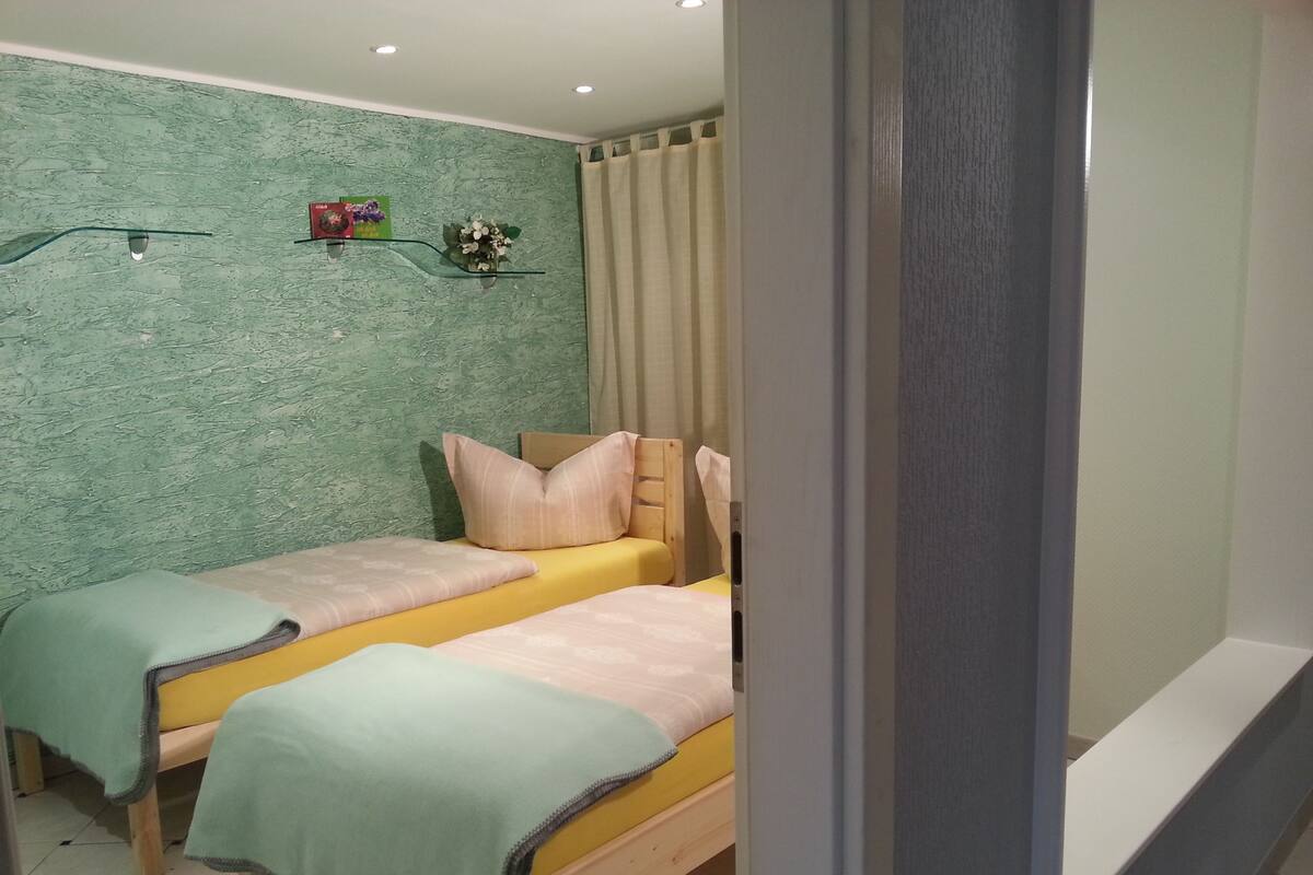 A bedroom with two single beds, a green textured wall, a small shelf with decorative items, and a curtain.