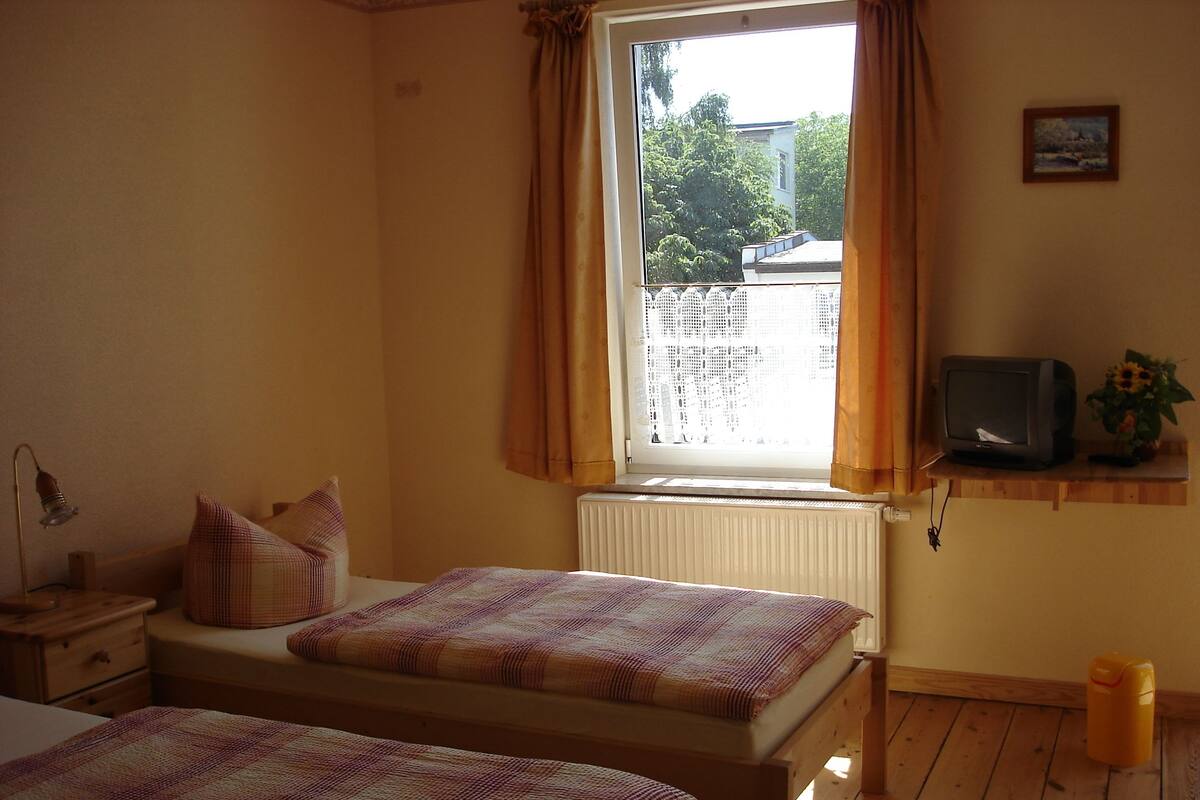 A bedroom with two beds, a window with curtains, a radiator, a television on a shelf, and a small table with a lamp.