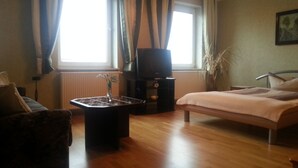 Ein Zimmer mit einem Bett, einem Fernseher auf einem Schrank, einem kleinen Tisch mit einer Vase und einem Fenster mit Vorhängen.