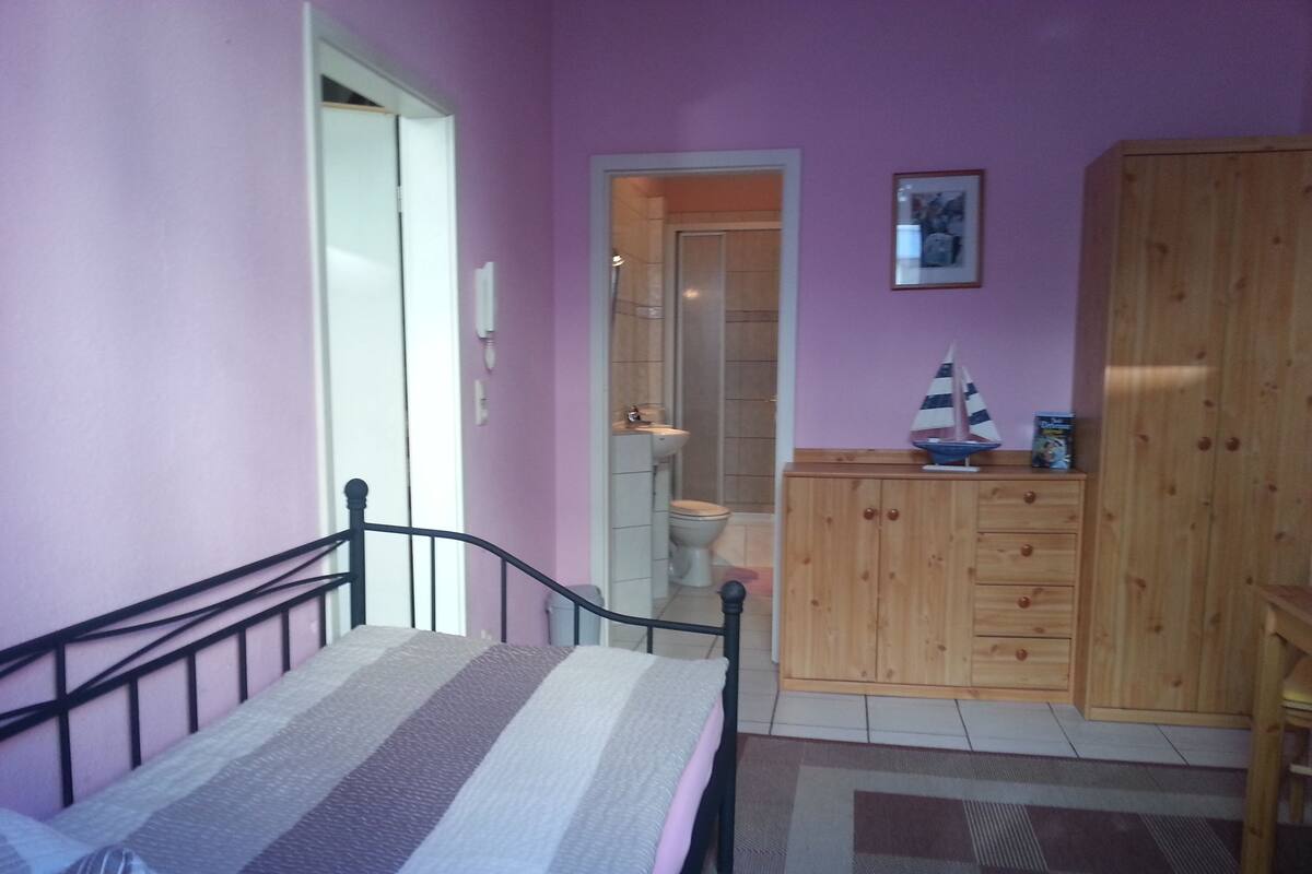 A bedroom with a bed, a wooden wardrobe, a small cabinet, and a door leading to a bathroom.