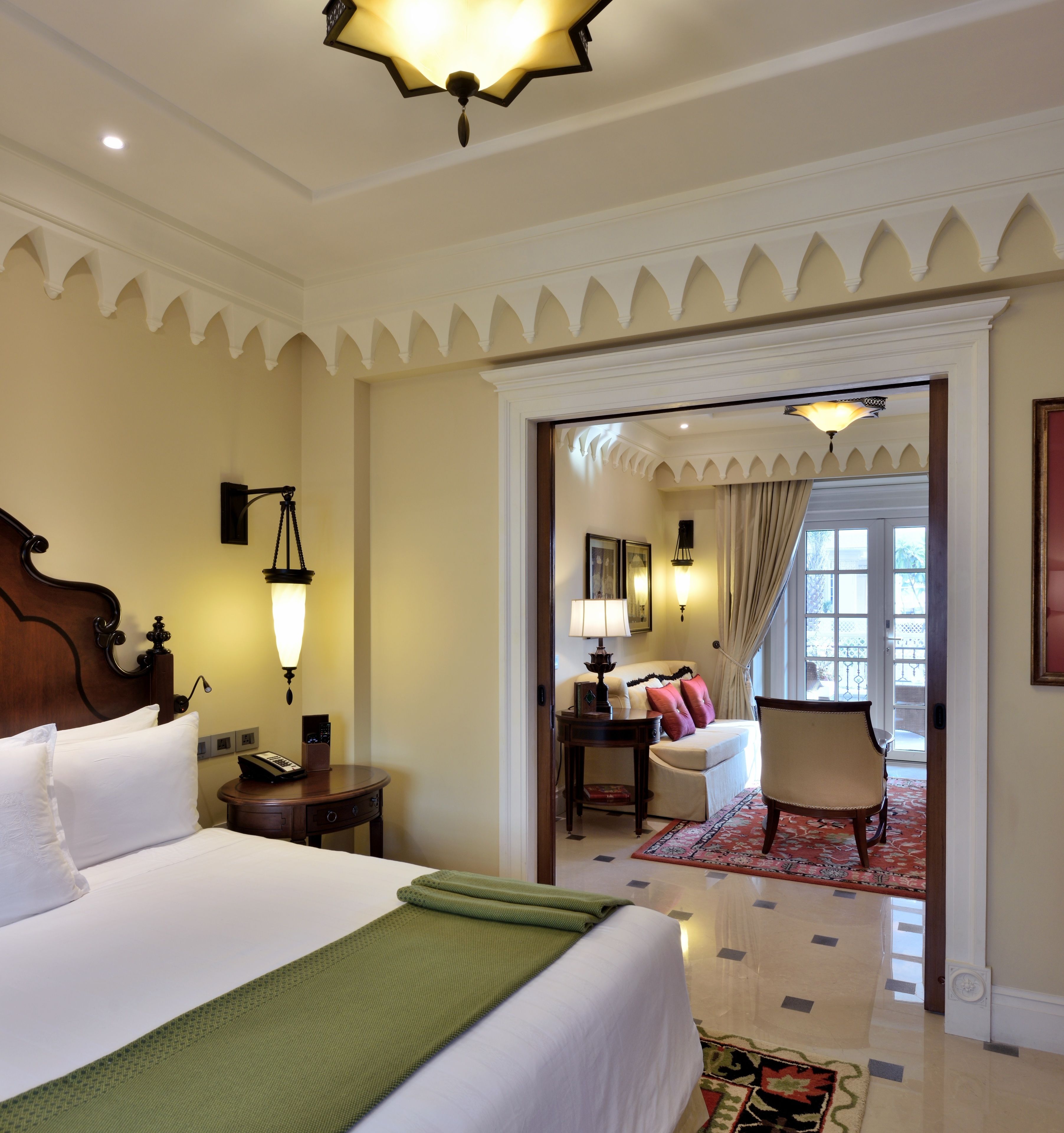 A hotel room with a large bed, a desk with a lamp, a chair, and a view through an open door to another room.
