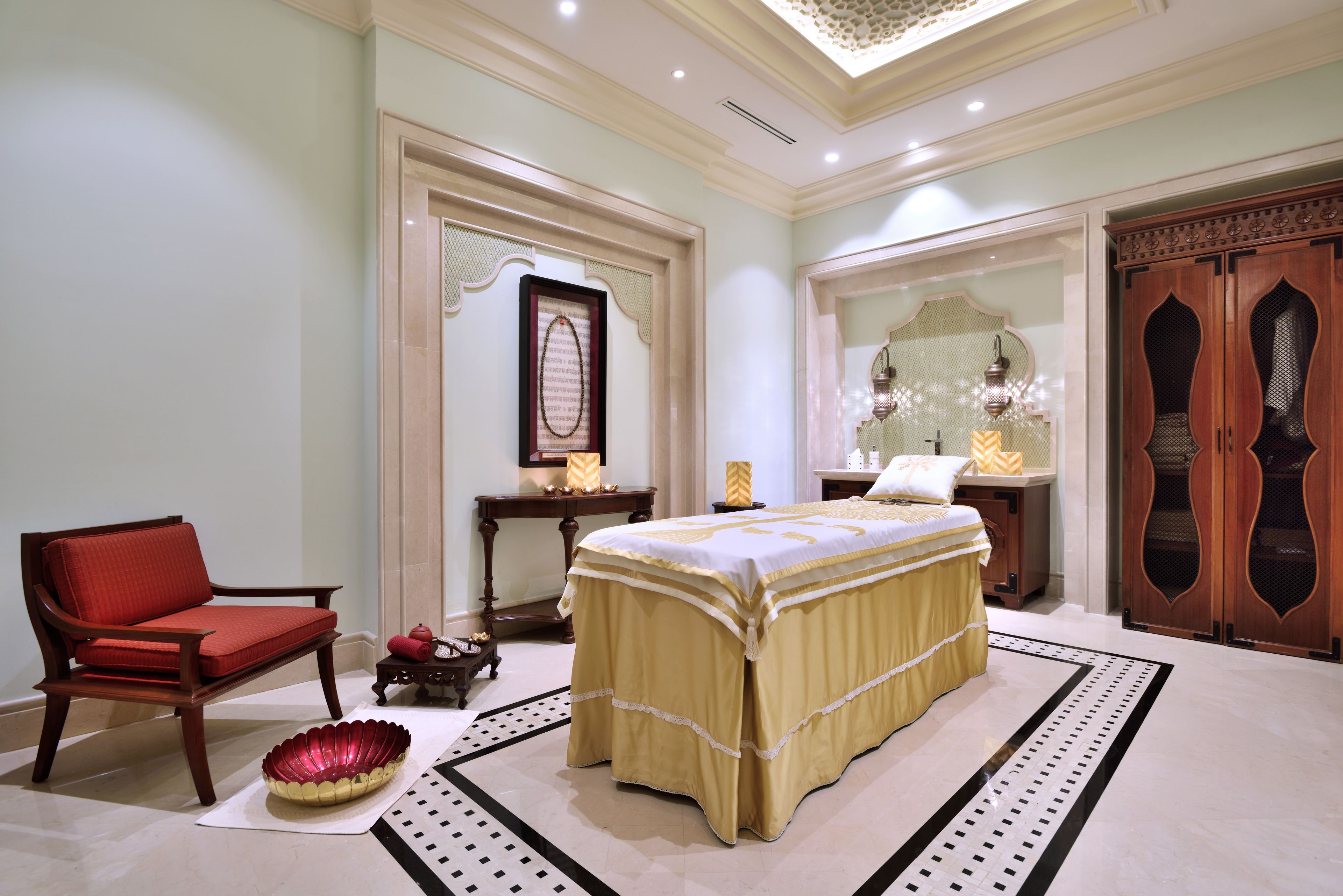 A room with a bed, a red chair, a small table with a lamp, and a decorative wall piece.