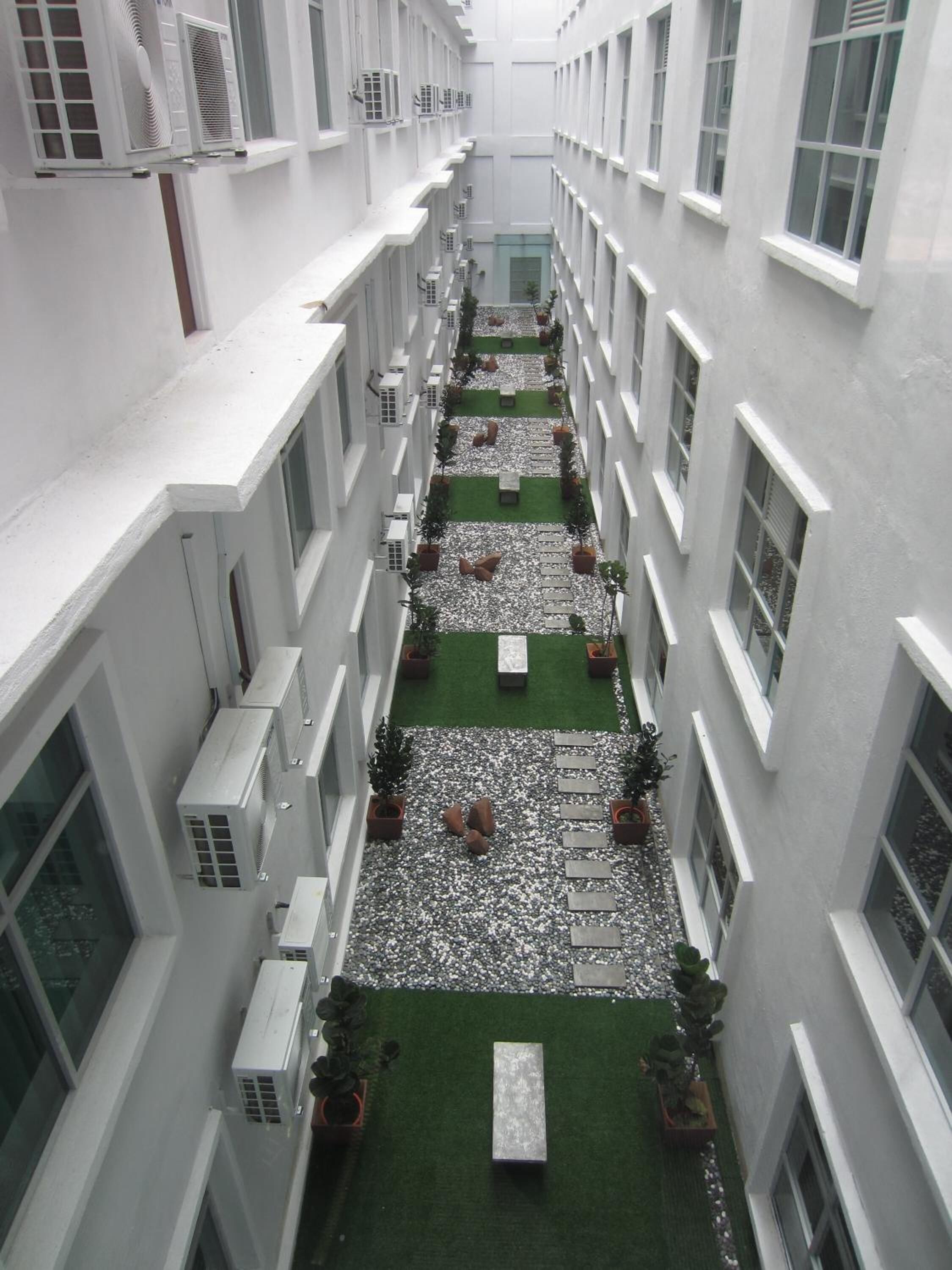 A narrow corridor with white walls, multiple windows, and air conditioning units. There are potted plants and a small green area with a bench.