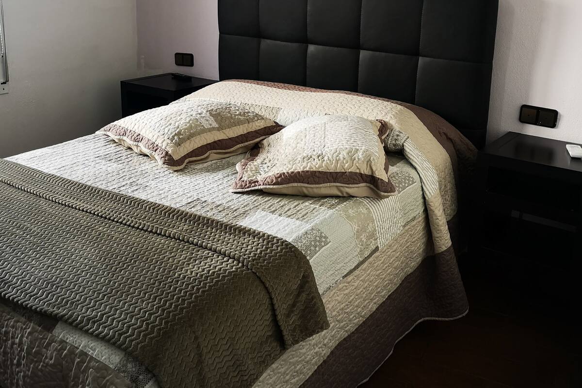 A neatly made bed with a quilted cover and two pillows, flanked by two bedside tables with a remote control on one.
