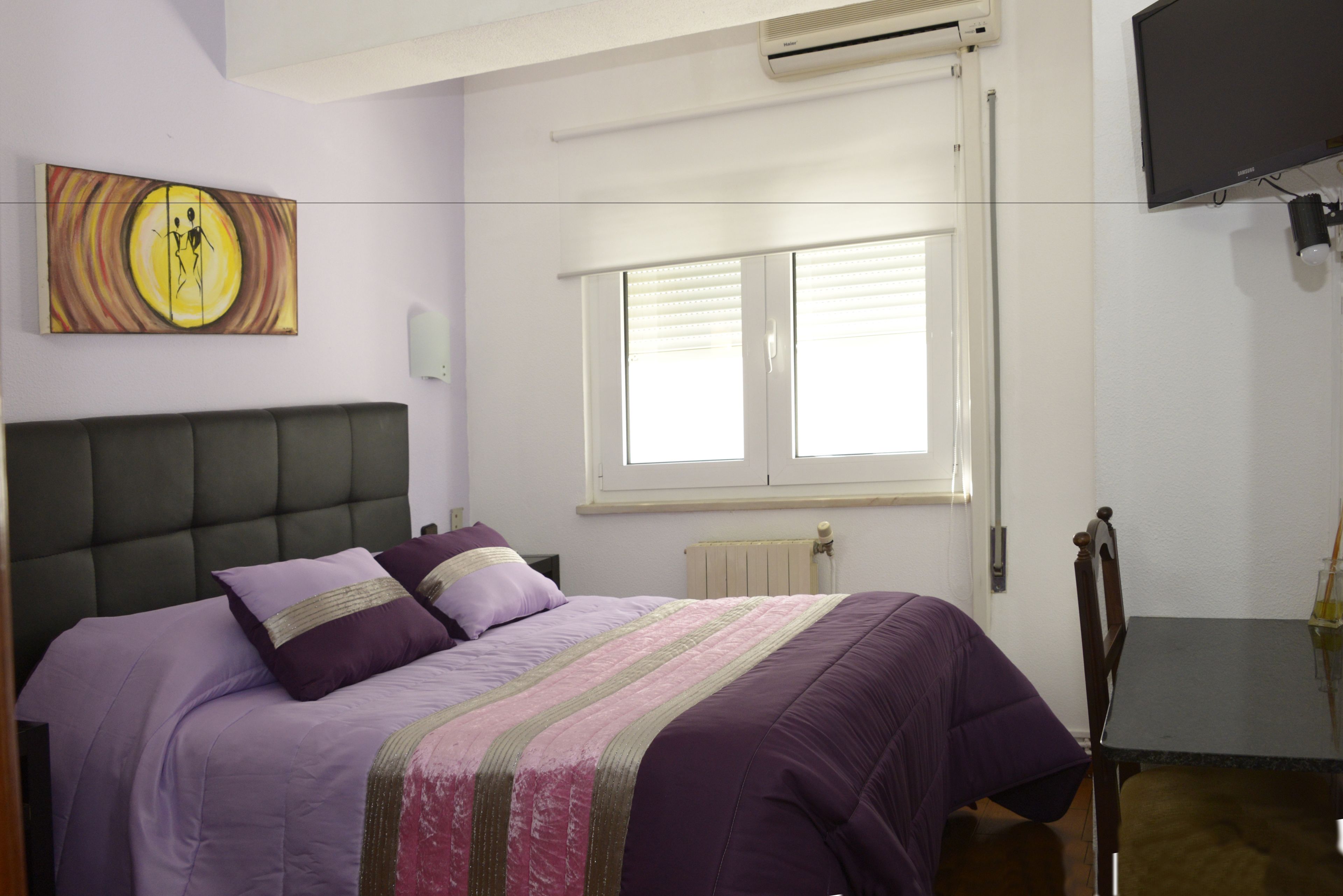 A bedroom with a bed, a window with blinds, a wall-mounted TV, and a radiator.