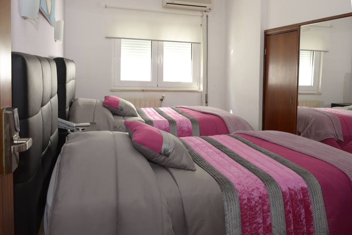 A hotel room with two beds, a wooden wardrobe, a radiator, and a window with blinds.