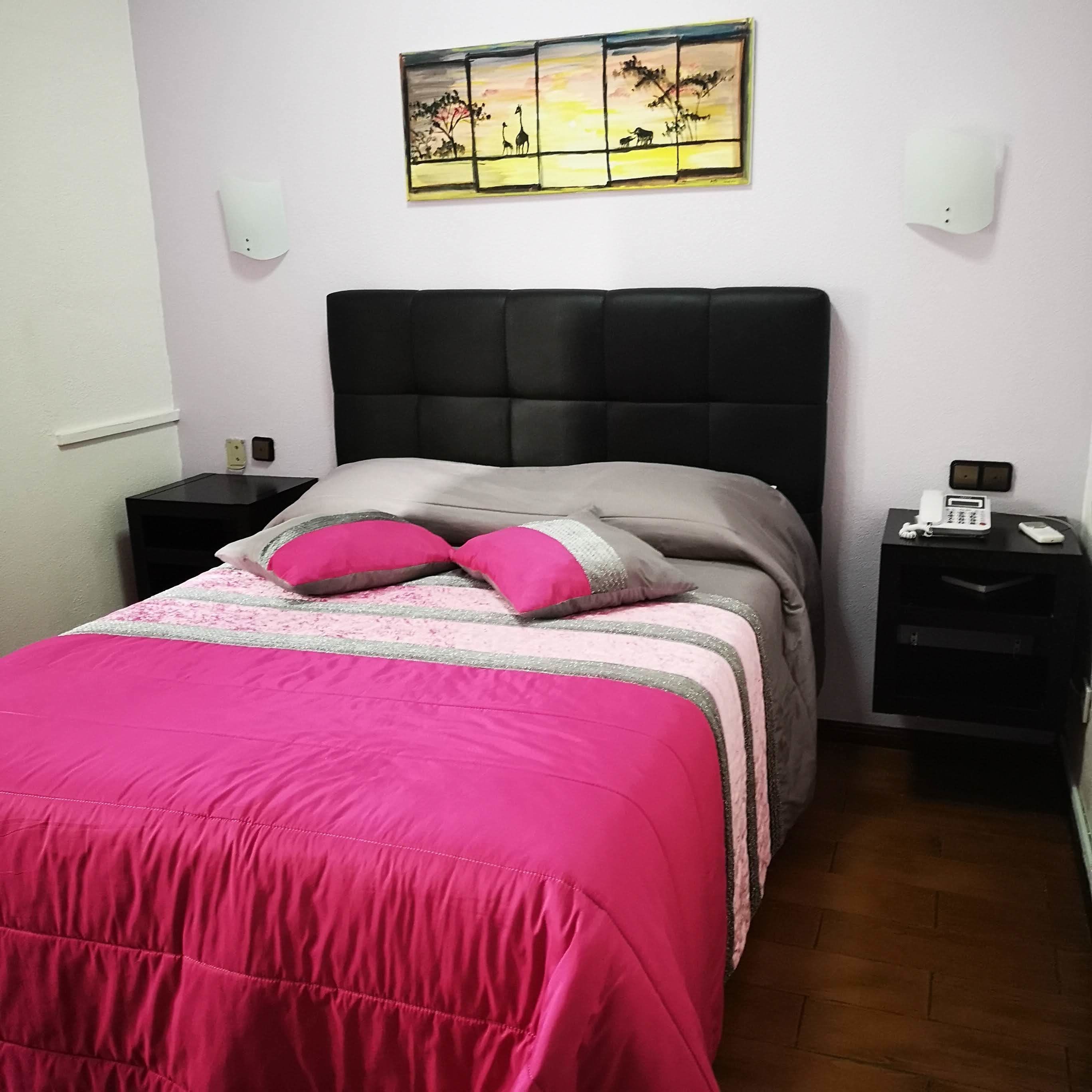 A bedroom with a black headboard, a pink bedspread, and two pink pillows. There is a painting on the wall above the bed, a nightstand with a phone, and a small table with a lamp.