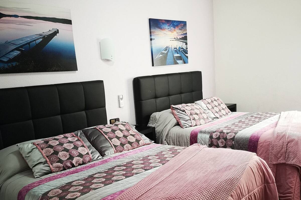 A hotel room with two beds, each with a headboard and pillows. There are wall-mounted light fixtures and a painting above the beds.