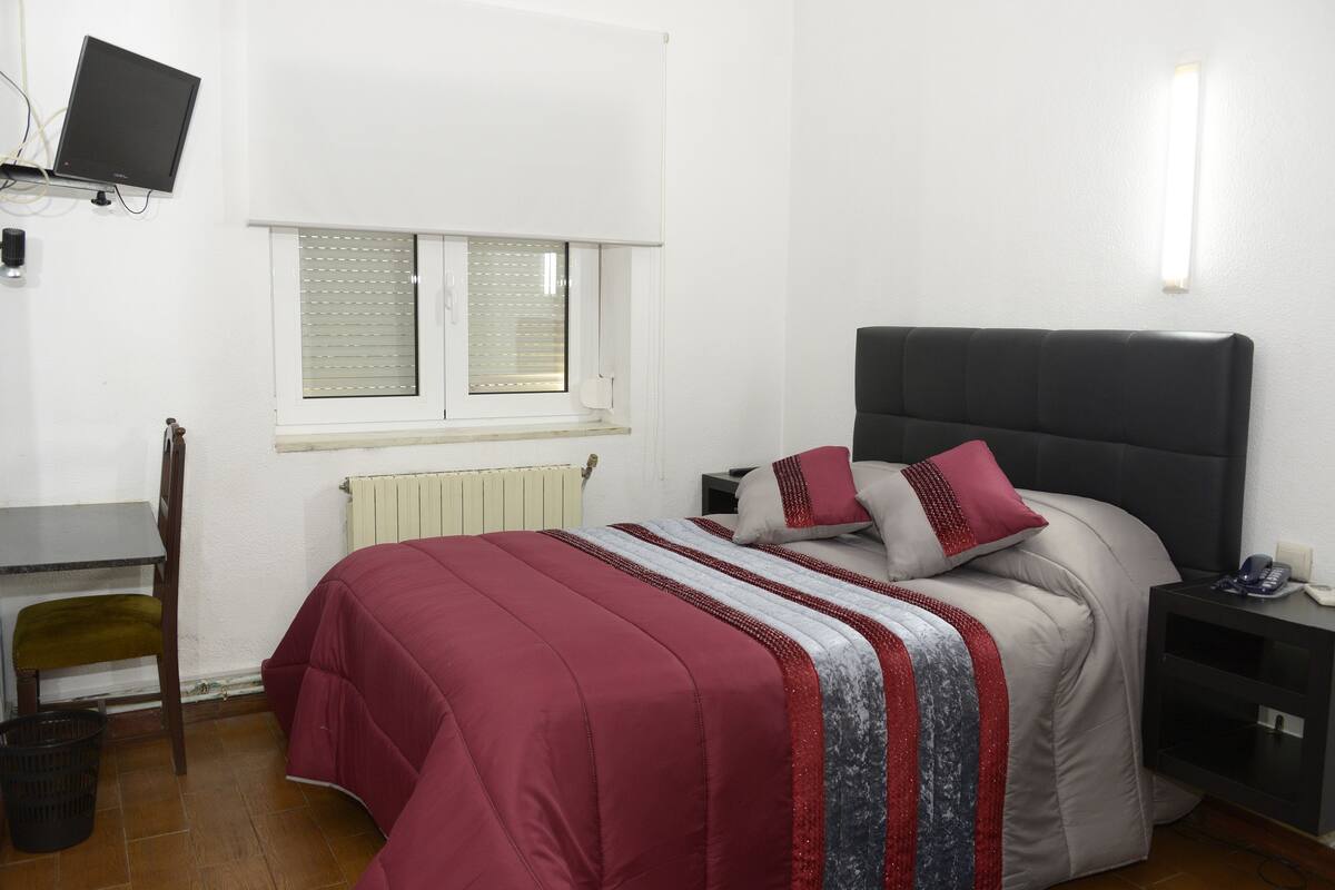 A bedroom with a bed, a desk, a chair, a window, and a nightstand.