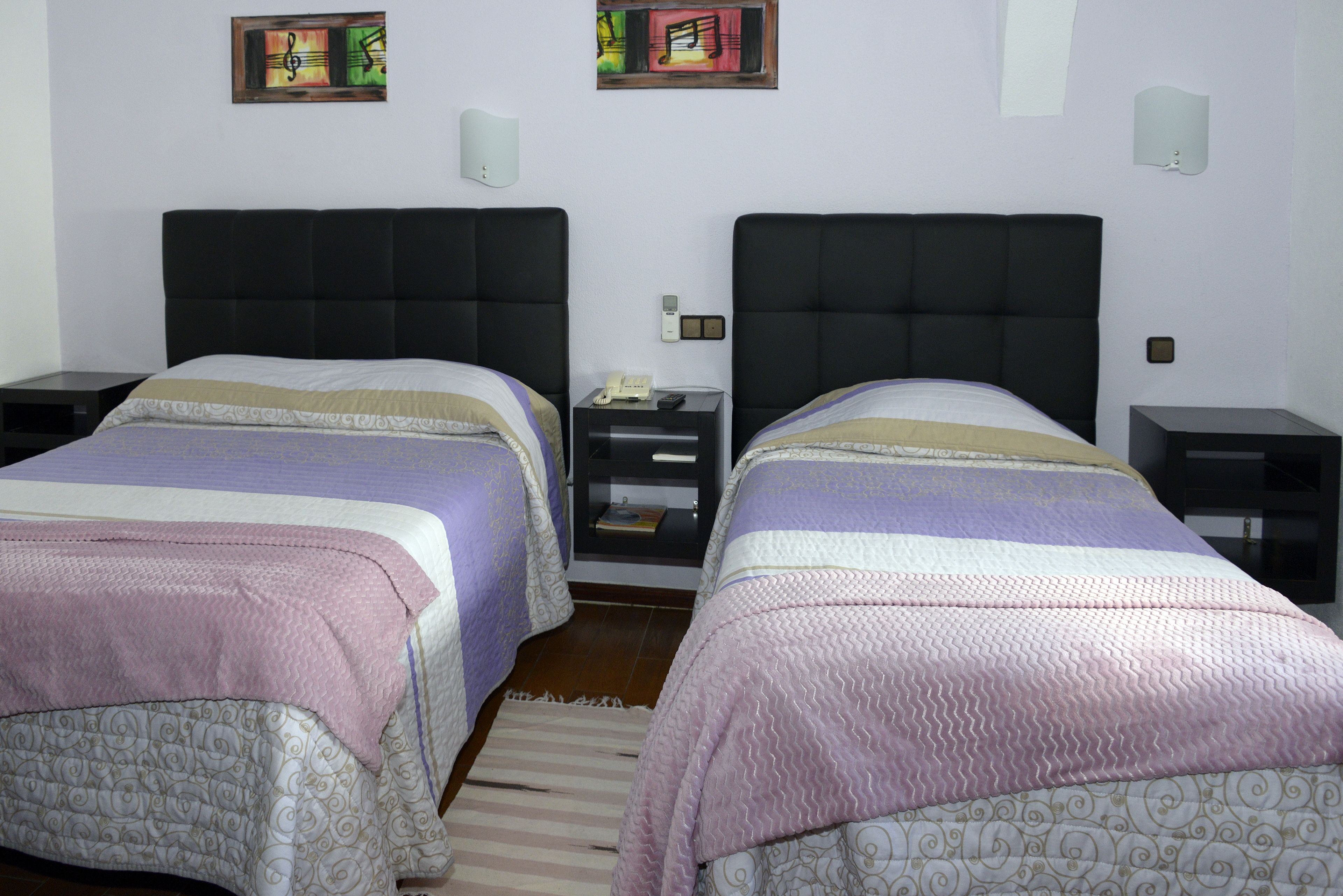 A hotel room with two beds, each with a headboard, a bedside table, and a nightstand. There are wall-mounted light fixtures and a framed picture on the wall.