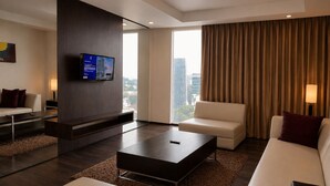 Een moderne hotelkamer met een flatscreen televisie aan een donkere muur, een groot raam met gordijnen, een witte bank, een bijzettafeltje en een kamerbreed tapijt.
