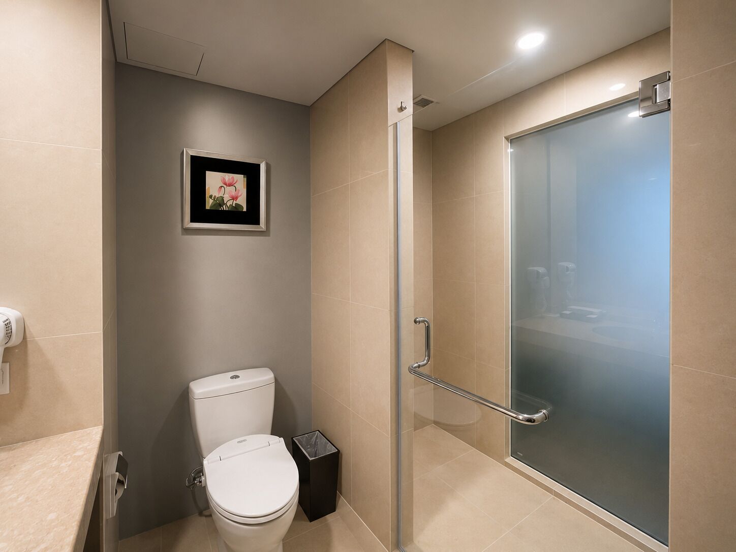 A modern bathroom with a toilet, a trash bin, a glass shower enclosure, and a framed picture on the wall.