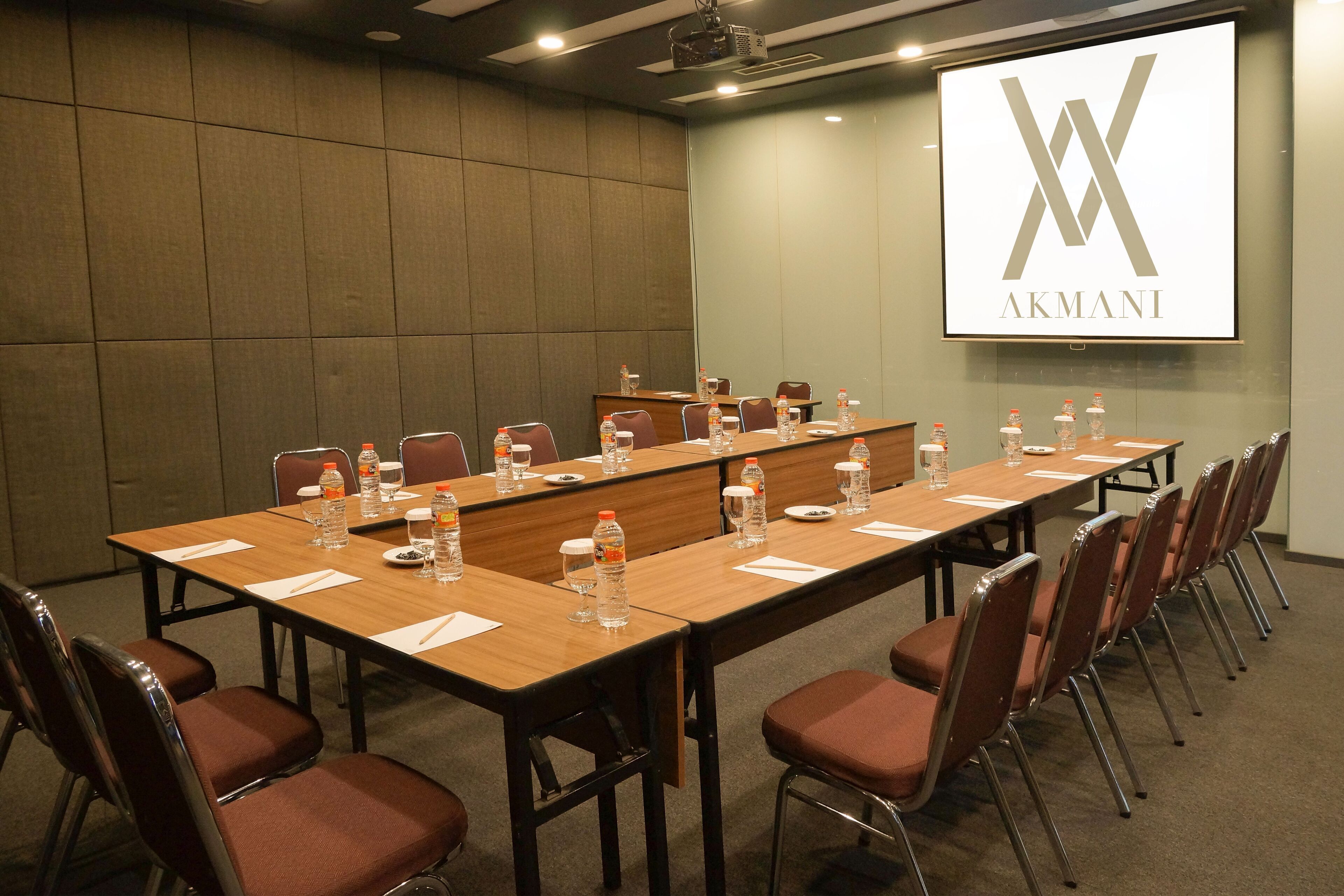 Un salón de conferencias con una larga mesa, sillas, botellas de agua y una pantalla de proyector mostrando el logotipo de AKMANI.