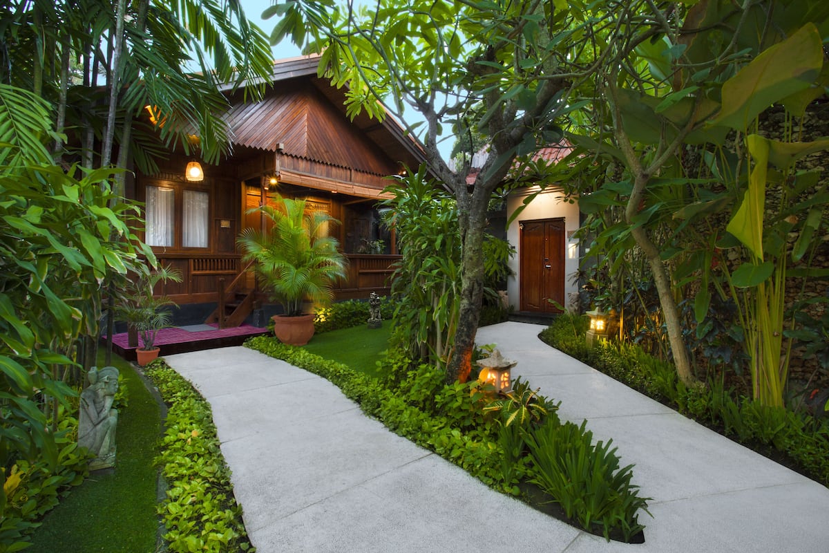 A wooden house with a porch, surrounded by lush greenery and a concrete pathway.