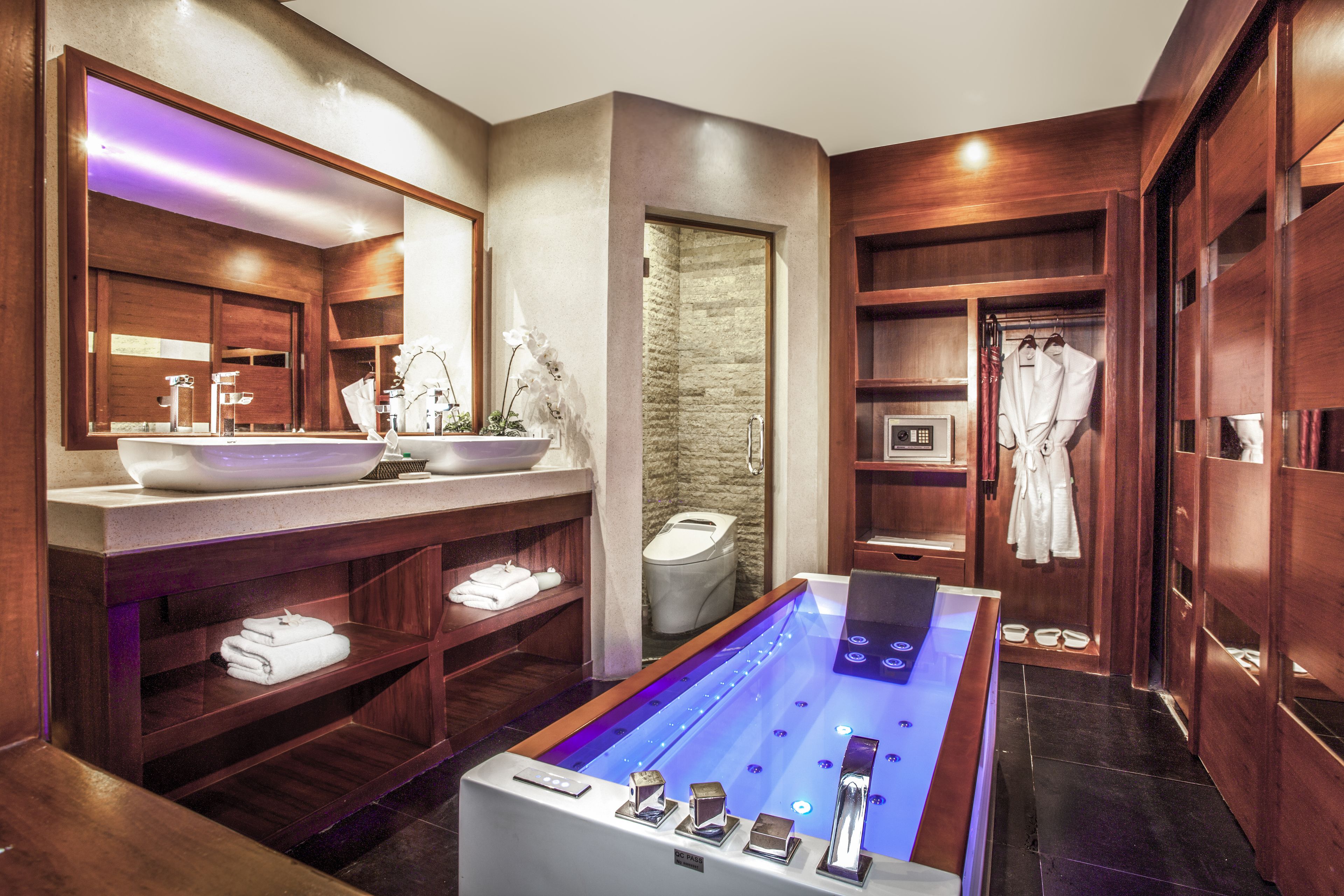 A modern bathroom with a large bathtub, wooden shelving, a vanity with a large mirror, and a stone wall.