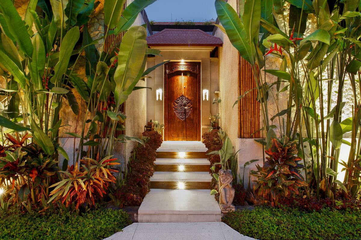 A well-lit entrance with a wooden door, steps leading up to it, and lush tropical plants on either side.