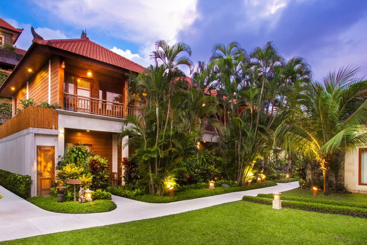 A two-story wooden house with a balcony, surrounded by lush tropical vegetation and a well-maintained garden.
