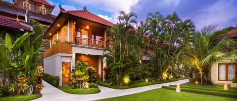 A two-story house with a balcony, surrounded by lush greenery and palm trees.