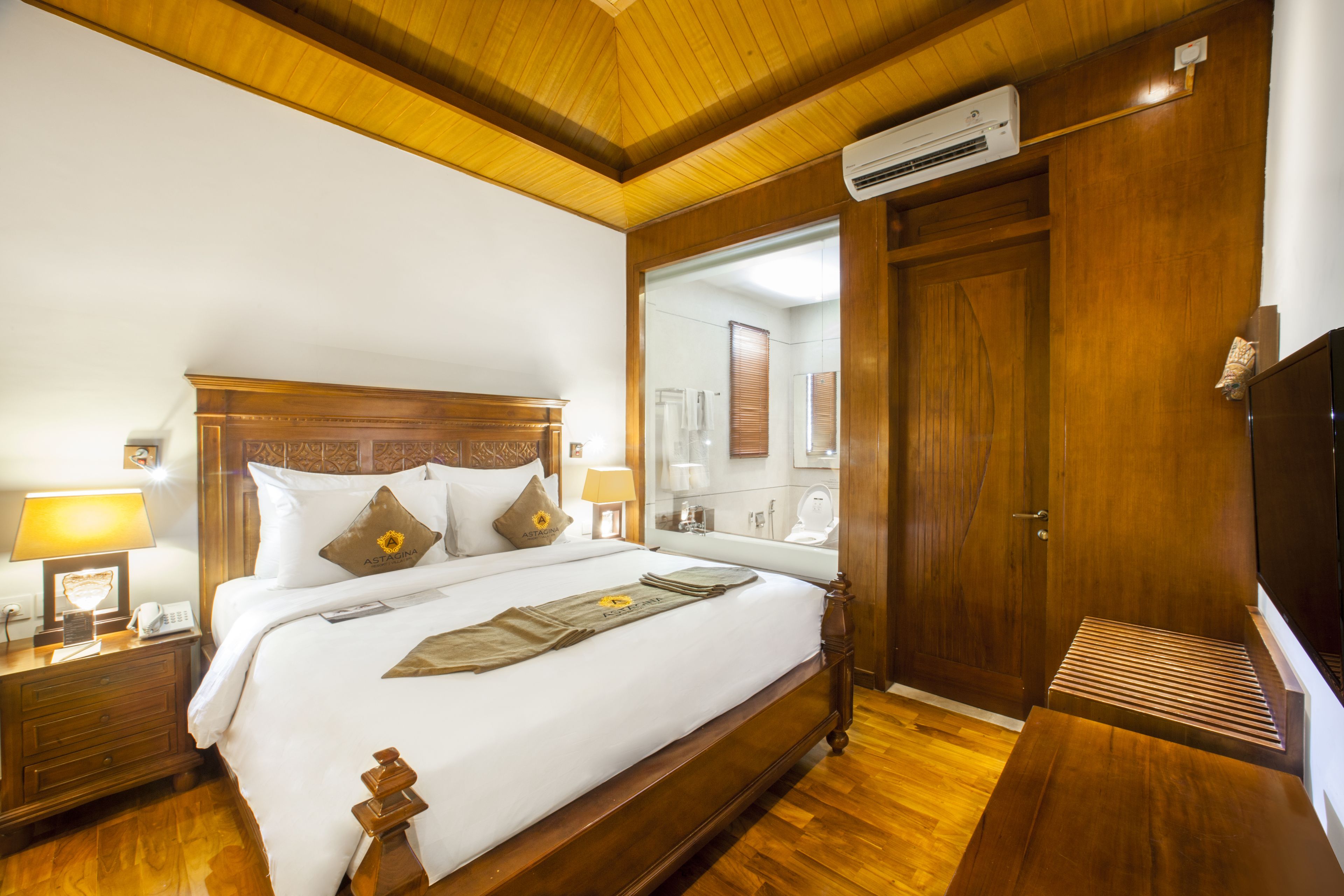 A hotel room with a wooden bed, a nightstand, a bedside lamp, a mirror, and a wooden bench.