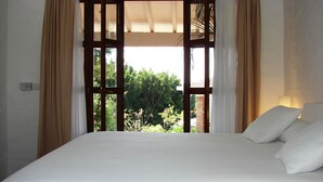 A bedroom with a large bed, beige curtains, and a view of greenery outside.