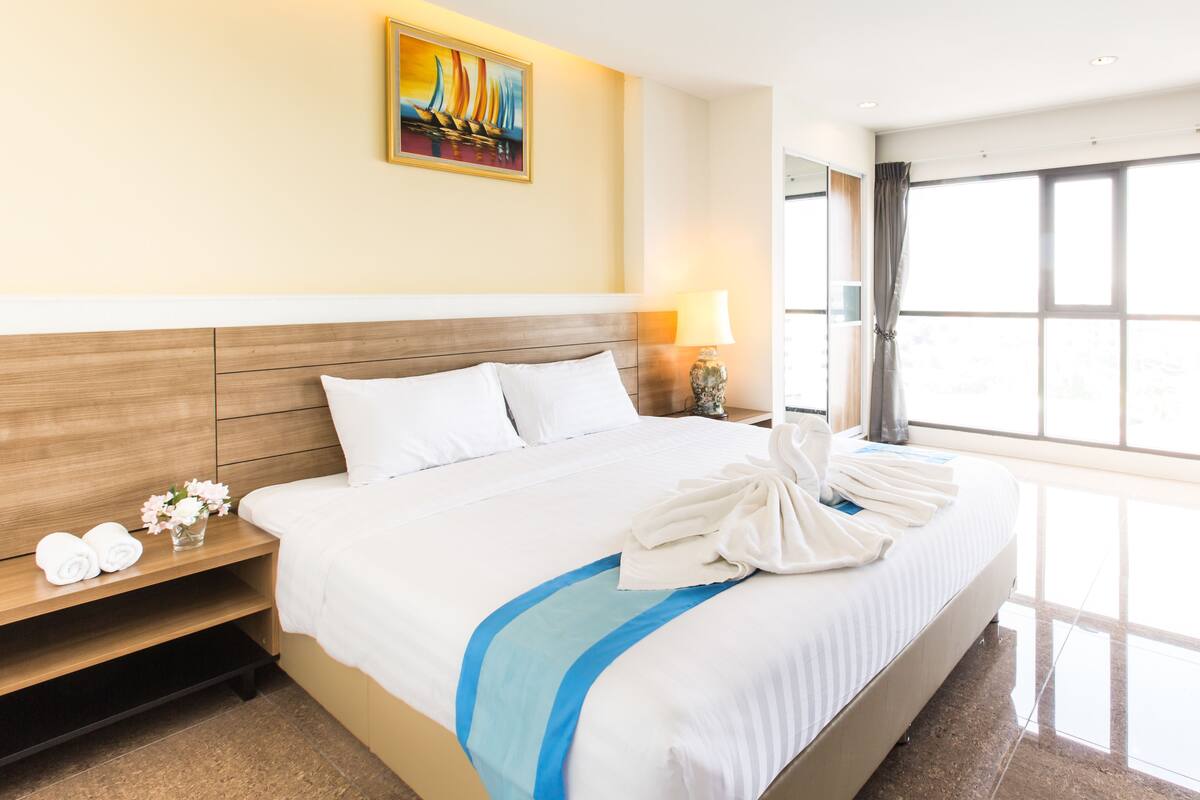 Habitación de hotel con una cama grande, una mesita de noche con lámpara, un cuadro en la pared y una ventana con cortinas.