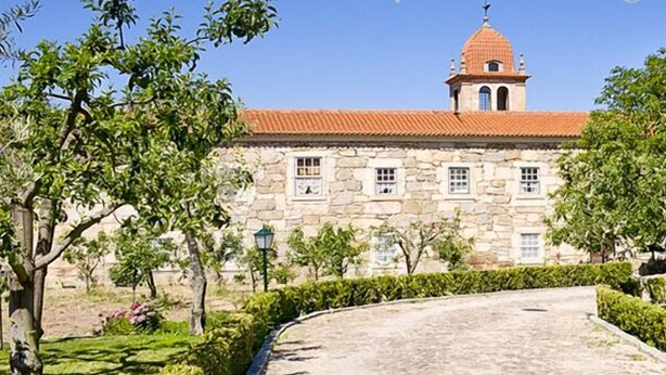 Um edifício de pedra com telhado de telhas vermelhas e uma torre de sino, rodeado por sebes bem cuidadas e árvores.