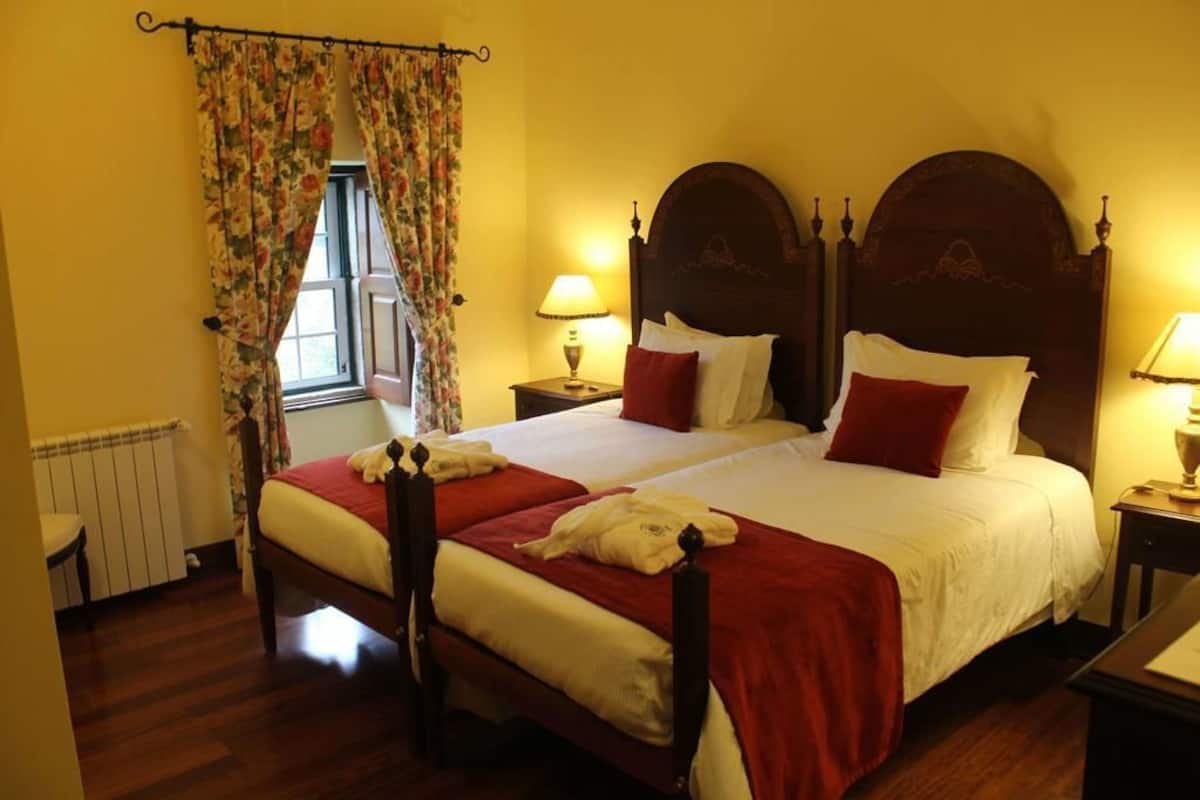 A double bed with a wooden headboard, two bedside tables with lamps, a window with floral curtains, and a radiator.
