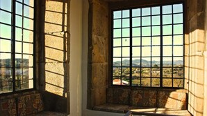 Quarto com uma grande janela que oferece vista para uma paisagem. Há um banco com almofadas estampadas.