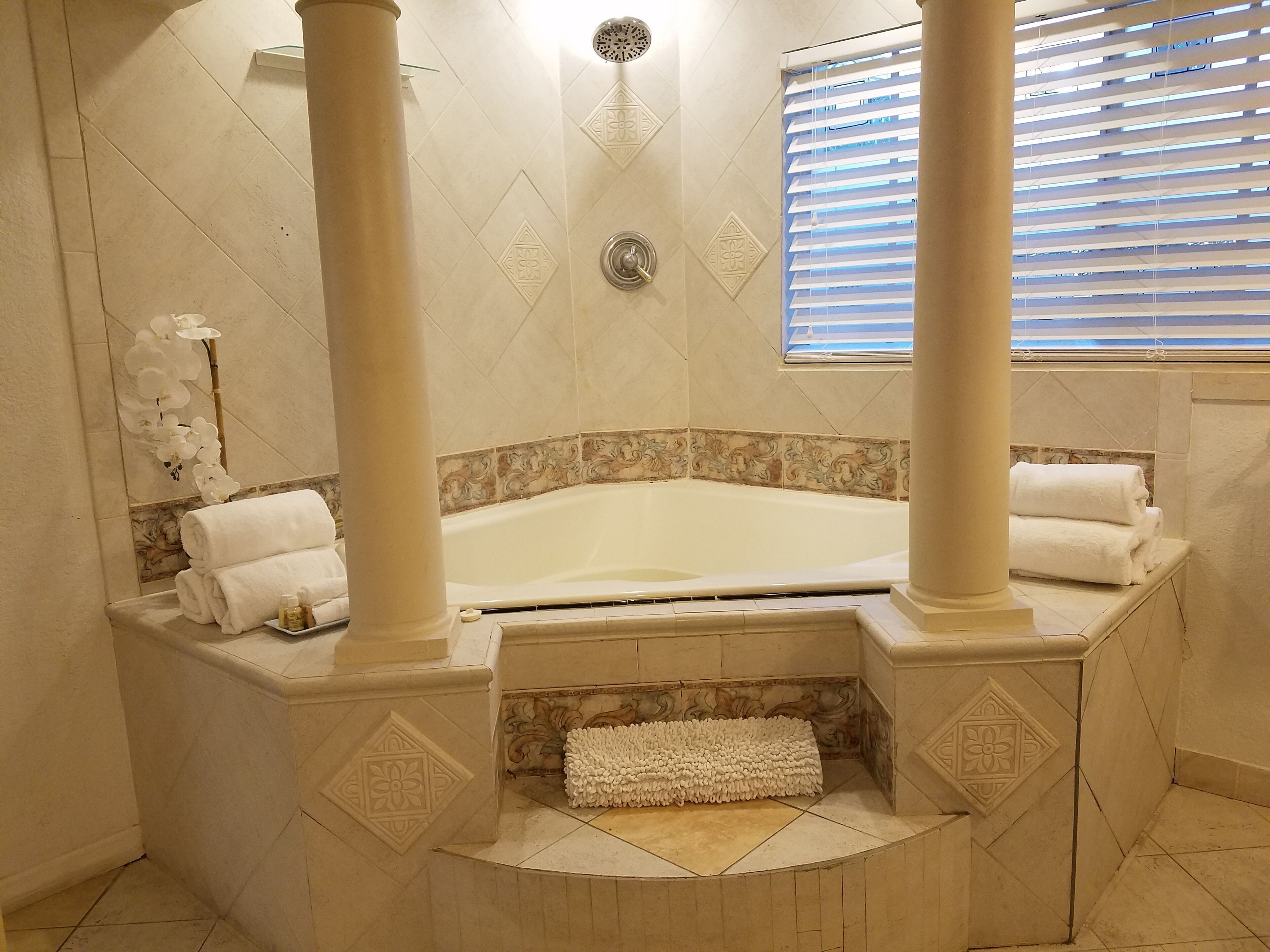 A bathroom with a bathtub, two columns, a window with blinds, and decorative tiles.