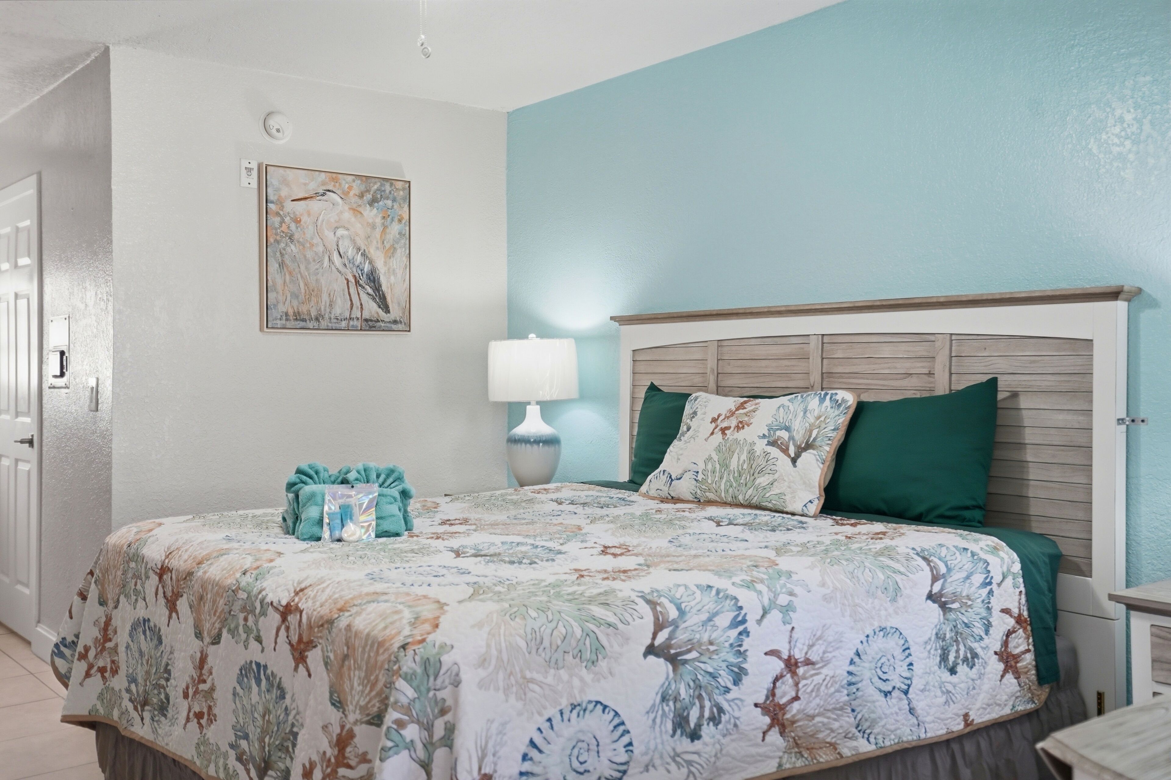 A bedroom with a floral bedspread, a wooden headboard, a bedside lamp, and a framed picture on the wall.