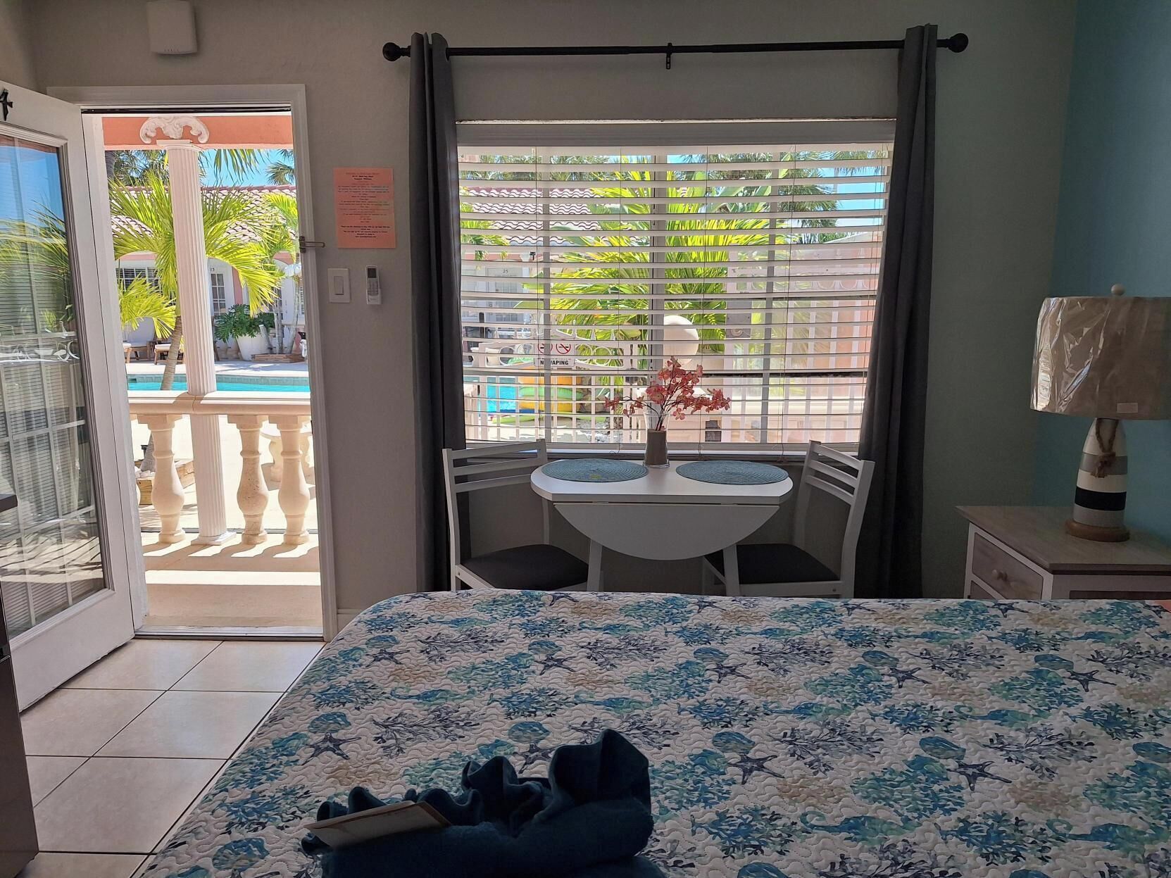 A hotel room with a bed, a small table with chairs, a window with blinds, and a view of a pool and palm trees.