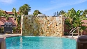 A swimming pool with a stone wall, a grill, and palm trees.