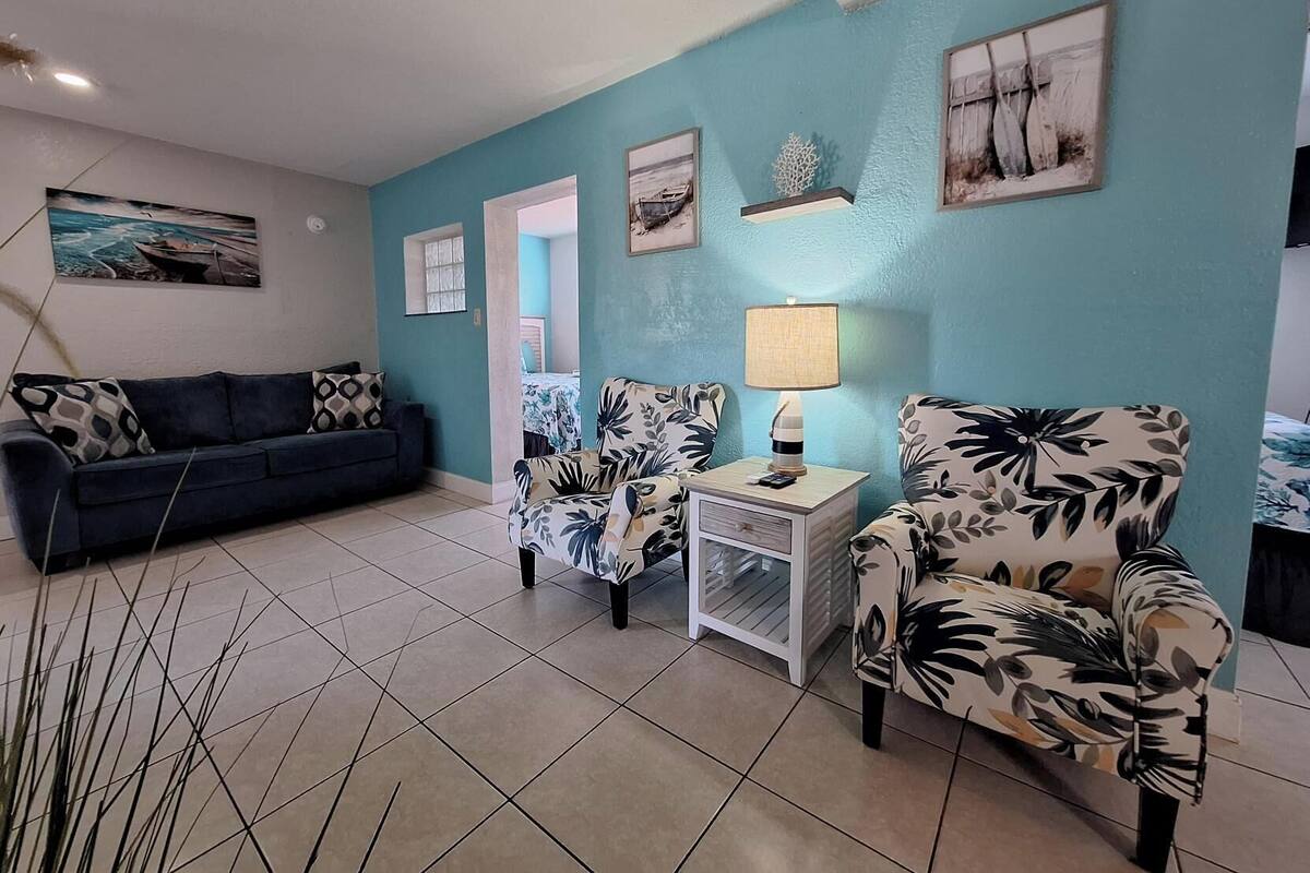 A living room with a blue wall, a sofa, two armchairs, a side table with a lamp, and a small shelf with decorative items.