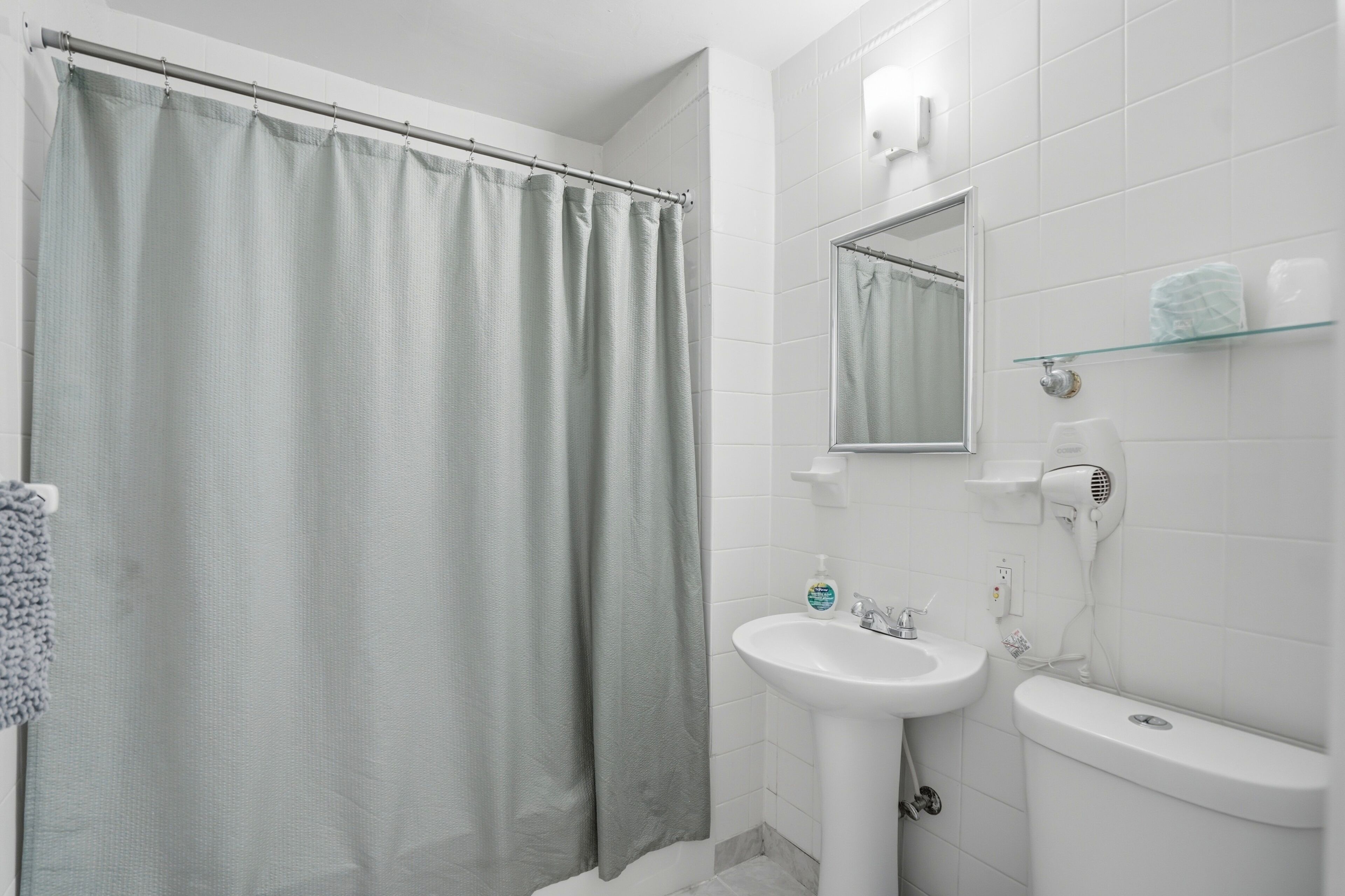 Ein Badezimmer mit Duschvorhang, Waschbecken, Spiegel, Ablagefläche aus Glas und einem Fön.