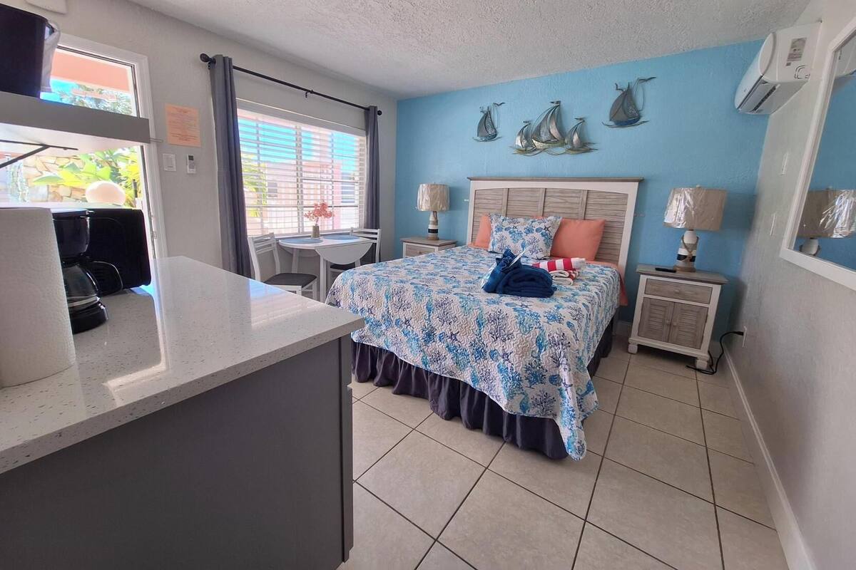 A bedroom with a bed, a small table, a chair, a coffee maker, and a window with curtains.