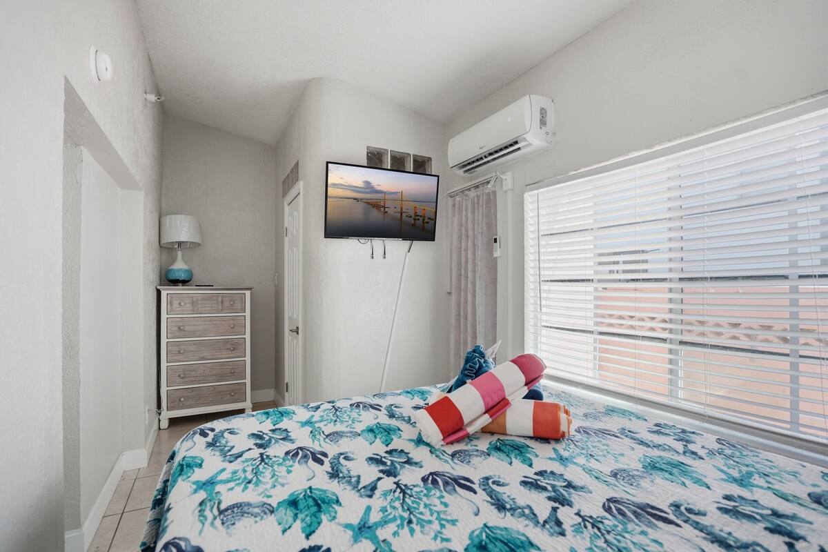 A bedroom with a bed, a TV mounted on the wall, a dresser, and a window with blinds.