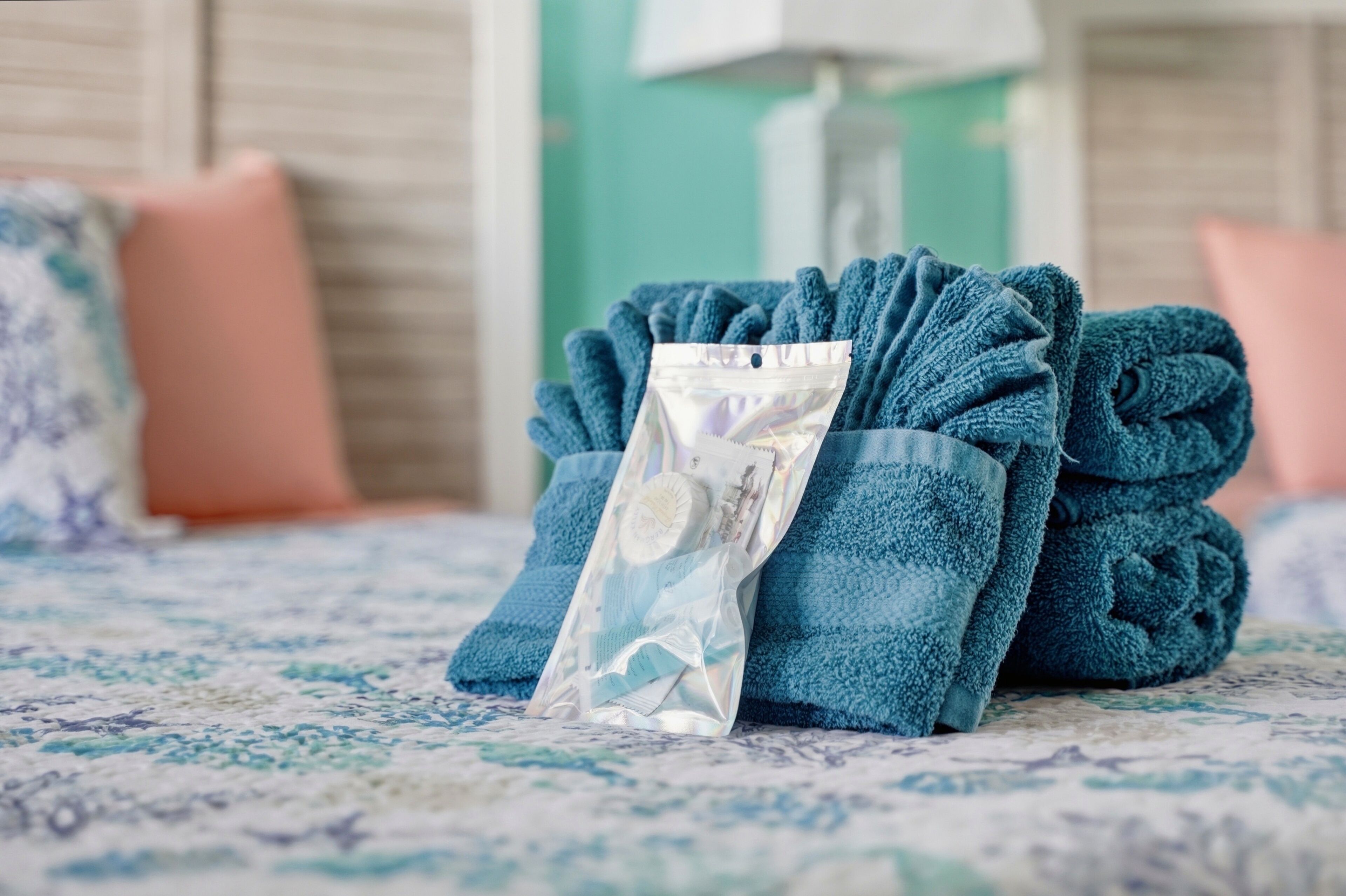 A neatly folded blue towel with a transparent plastic bag containing a bar of soap on a patterned bedspread.
