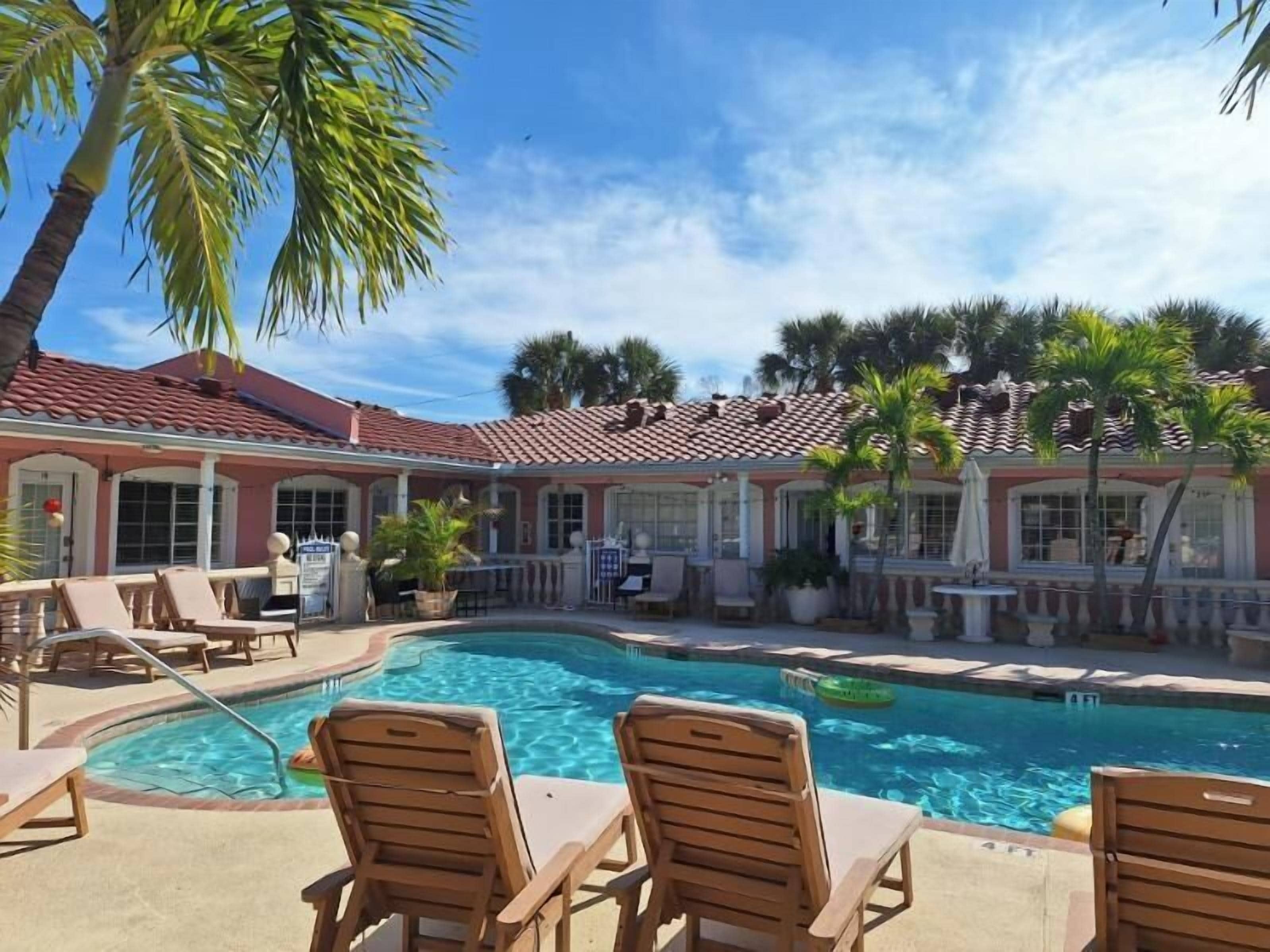 A pool with lounge chairs, a building with a tiled roof, and palm trees.