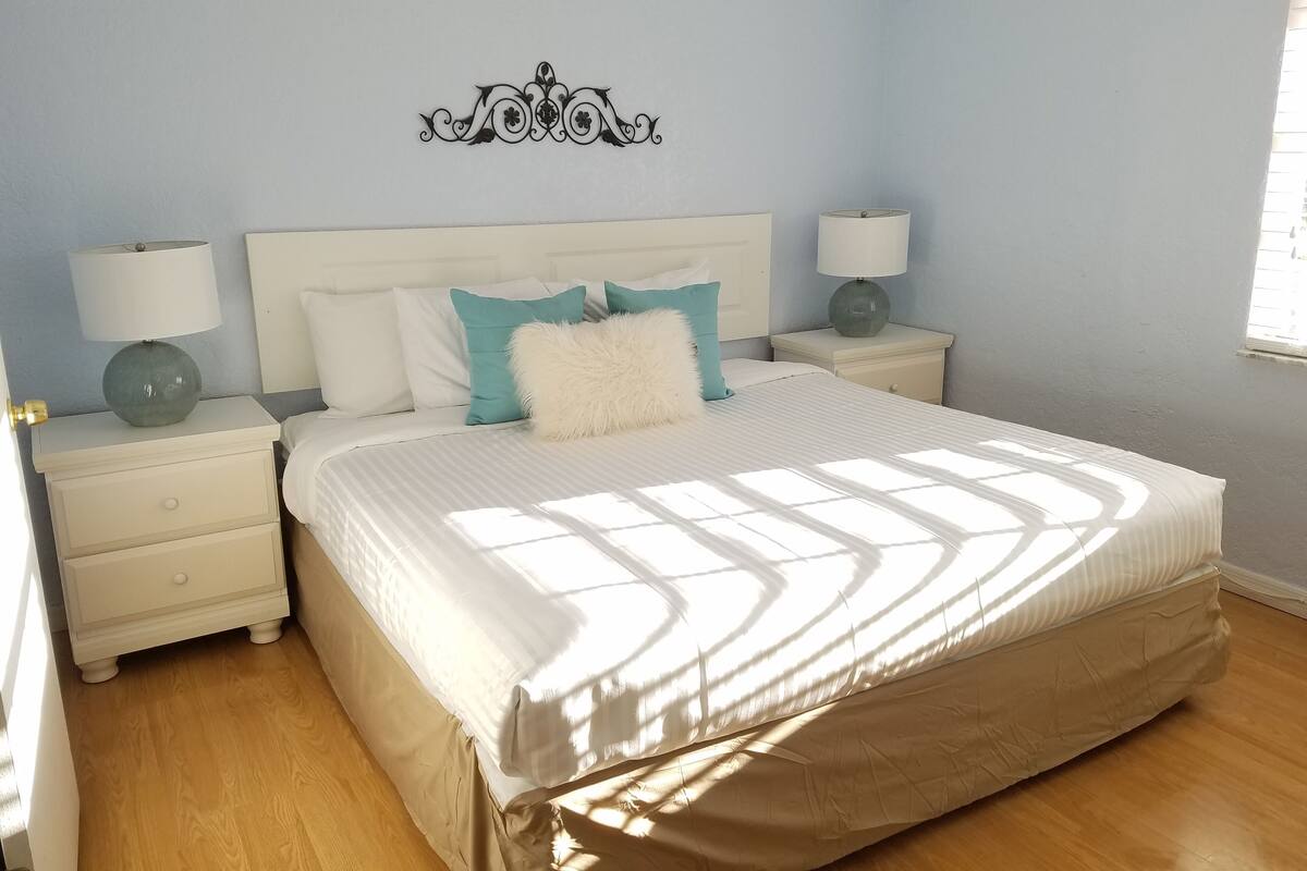 A bedroom with a large bed, two bedside tables with lamps, a decorative wall piece, and a window providing natural light.