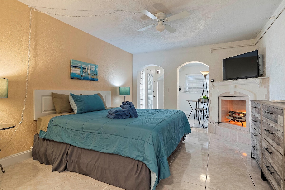 A bedroom with a bed, a television, a fireplace, and a hallway leading to another room.