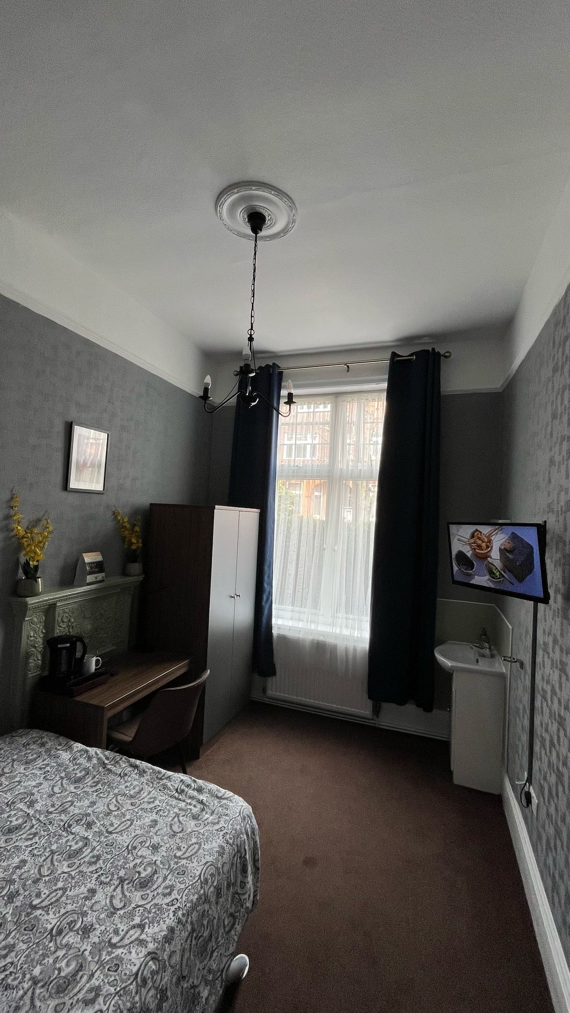 A bedroom with a bed, a desk, a chair, a wardrobe, a television, and a window with curtains.